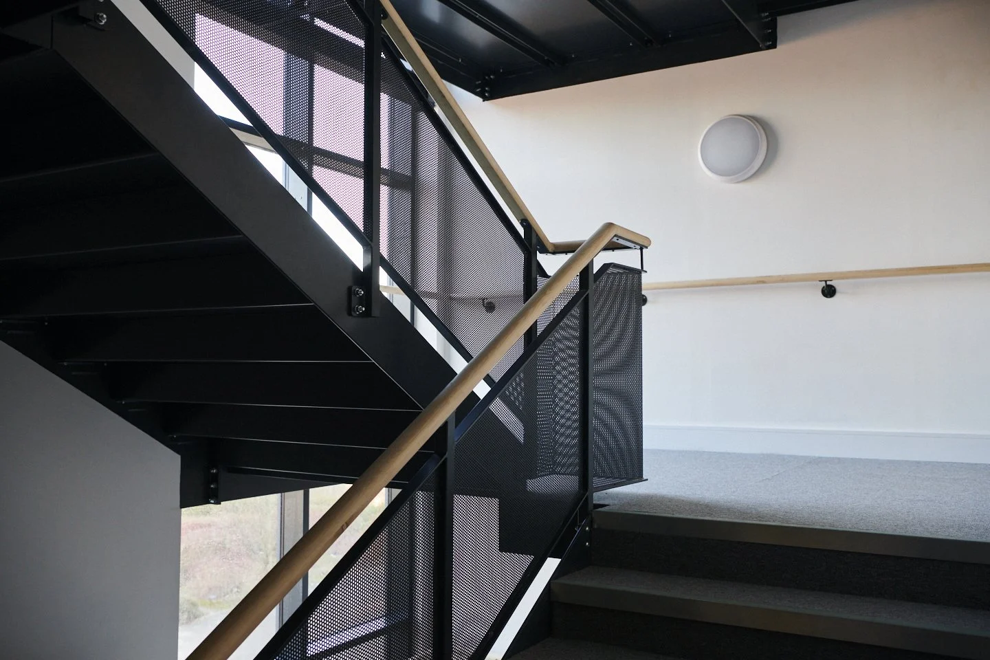 Interior view of a modern staircase with black metal railings, wooden handrails, and gray stairs in a well-lit space with white walls and a round ceiling light.