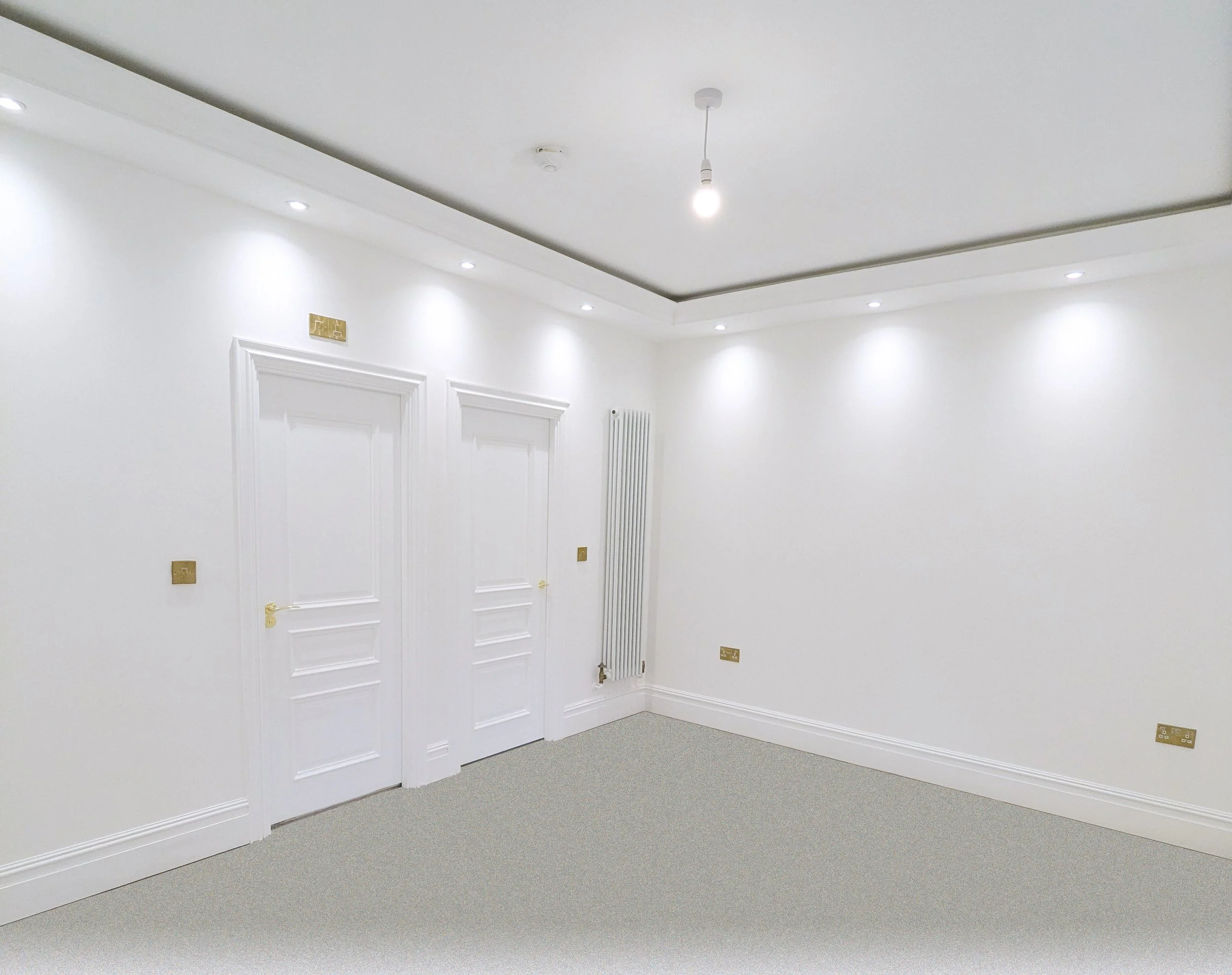 Empty white room with two closed white doors, gold handles, recessed ceiling lights, a white radiator, and beige carpet flooring.