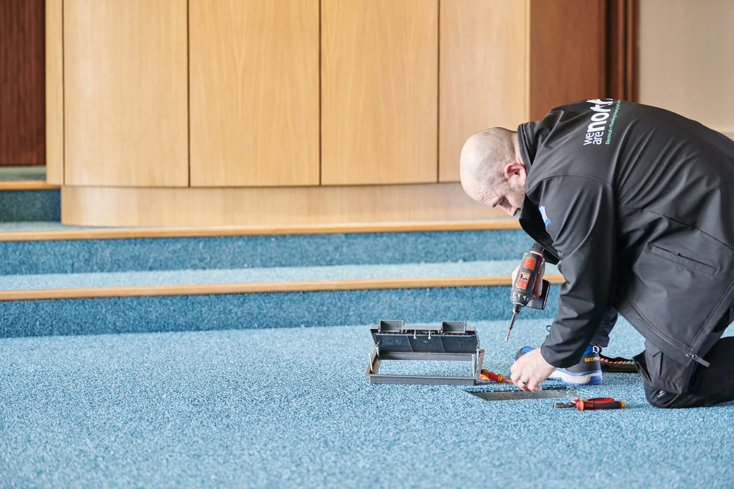 A man kneeling on blue carpeted stairs, installing or repairing an electrical outlet using a screwdriver and cordless drill.