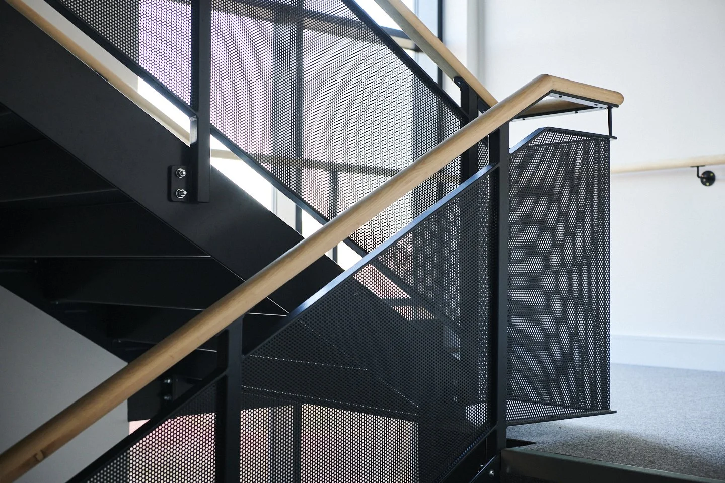Close-up view of a modern staircase with black metal perforated panels, light wood handrails, and black metal supports in a bright building interior.