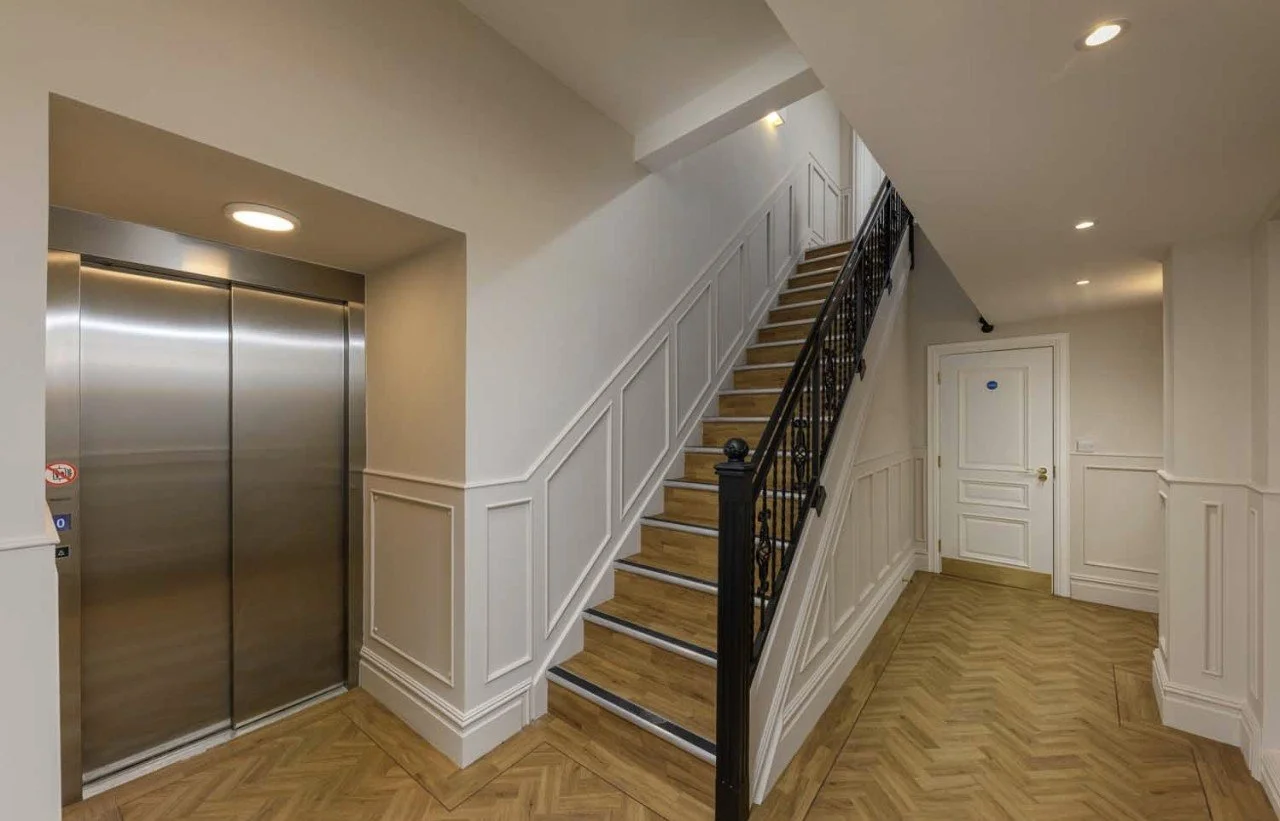 Interior view of a staircase with wooden steps and white paneling, an elevator with closed metal doors on the left, and a white door at the top of the stairs, with wood flooring and recessed ceiling lights.