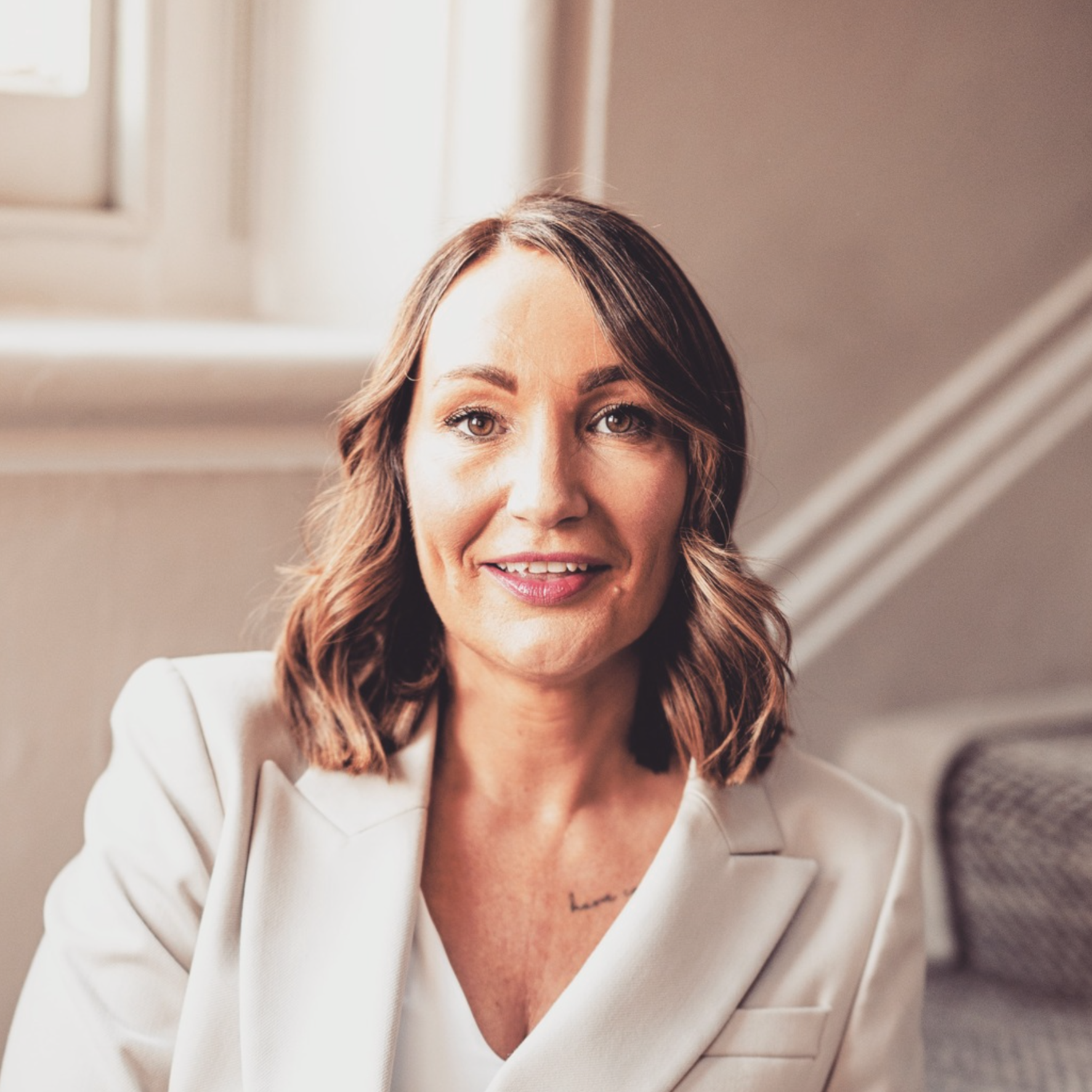 A woman with shoulder-length brown hair, smiling, sitting indoors near a window, wearing a white blazer.