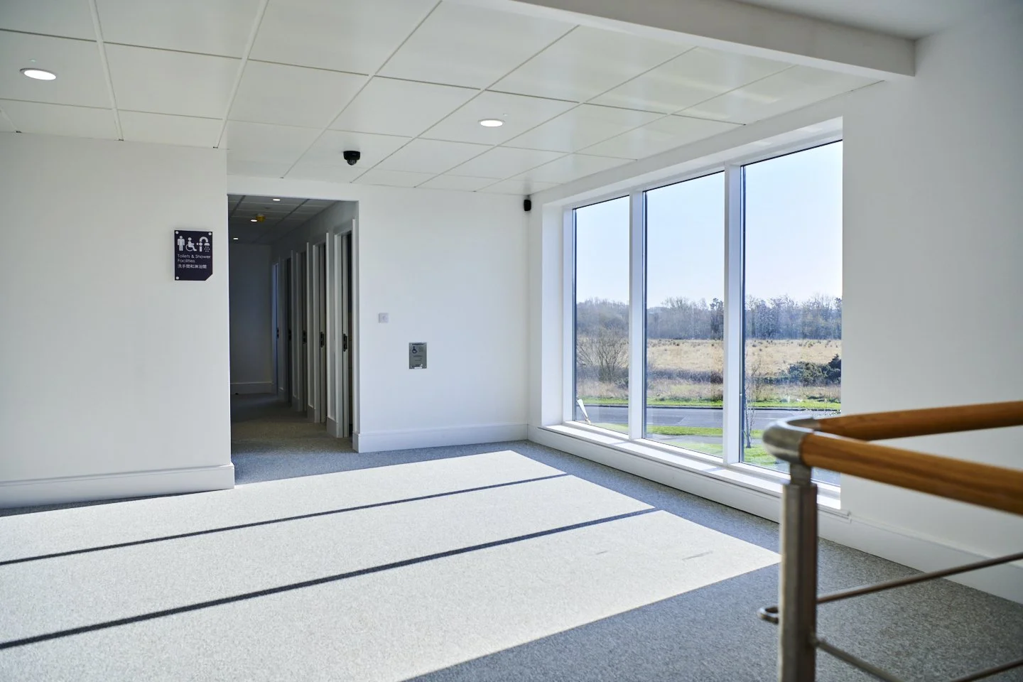 Bright, empty room with large windows showing outdoor scenery, overhead ceiling lights, and a wooden handrail in the foreground.