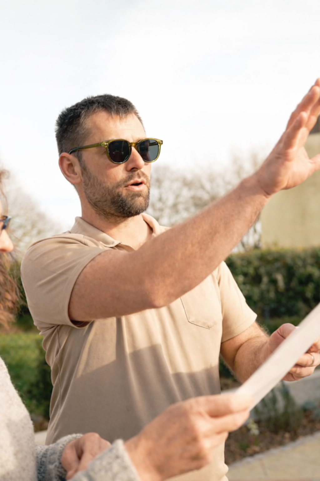 A man wearing sunglasses is outdoors, holding a garden design, and gesturing with his arm extended while talking.