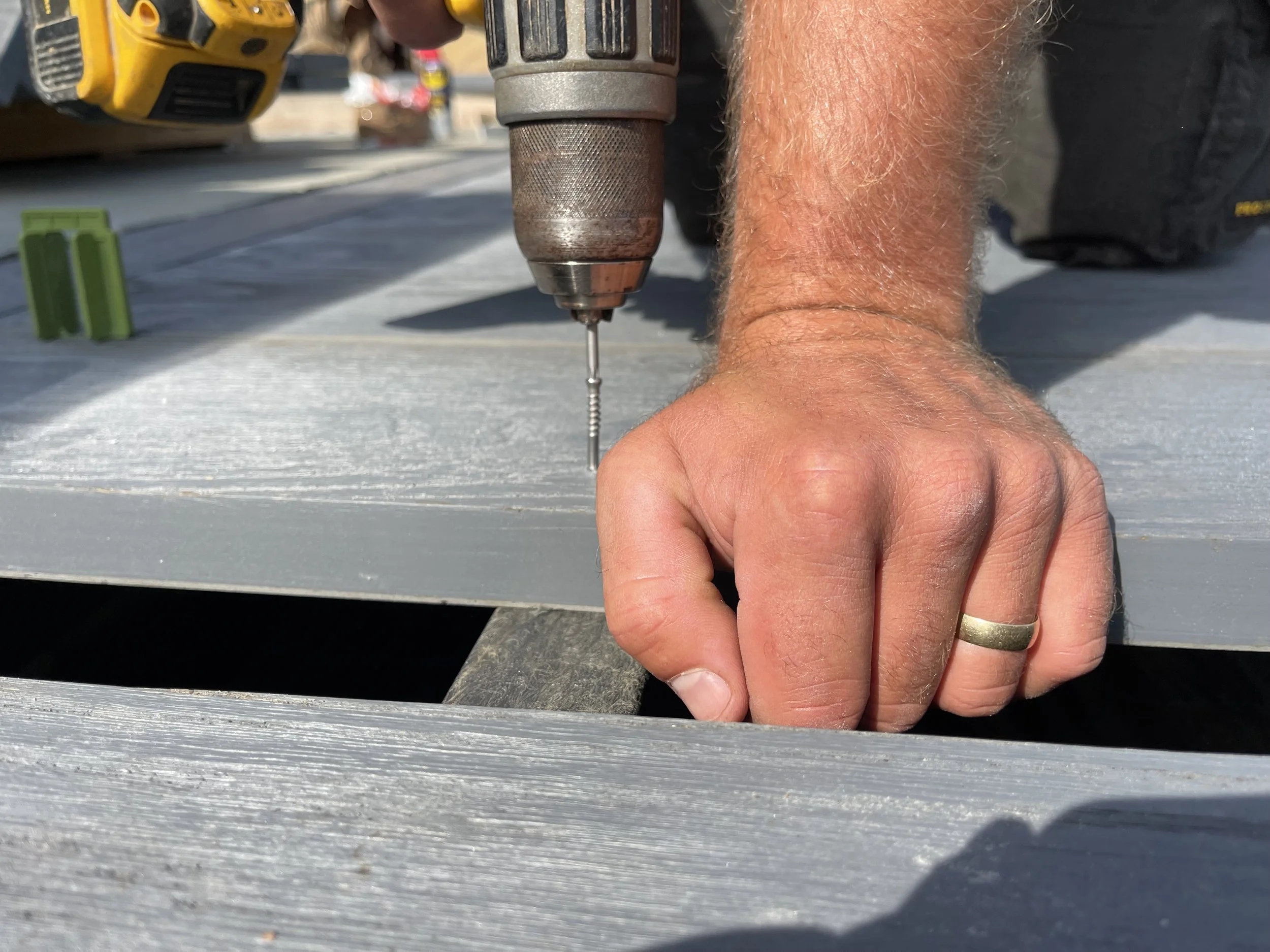 Close-up of a person's hand holding a silver screw while using a cordless drill to drive the screw into gray wooden planks outdoors.