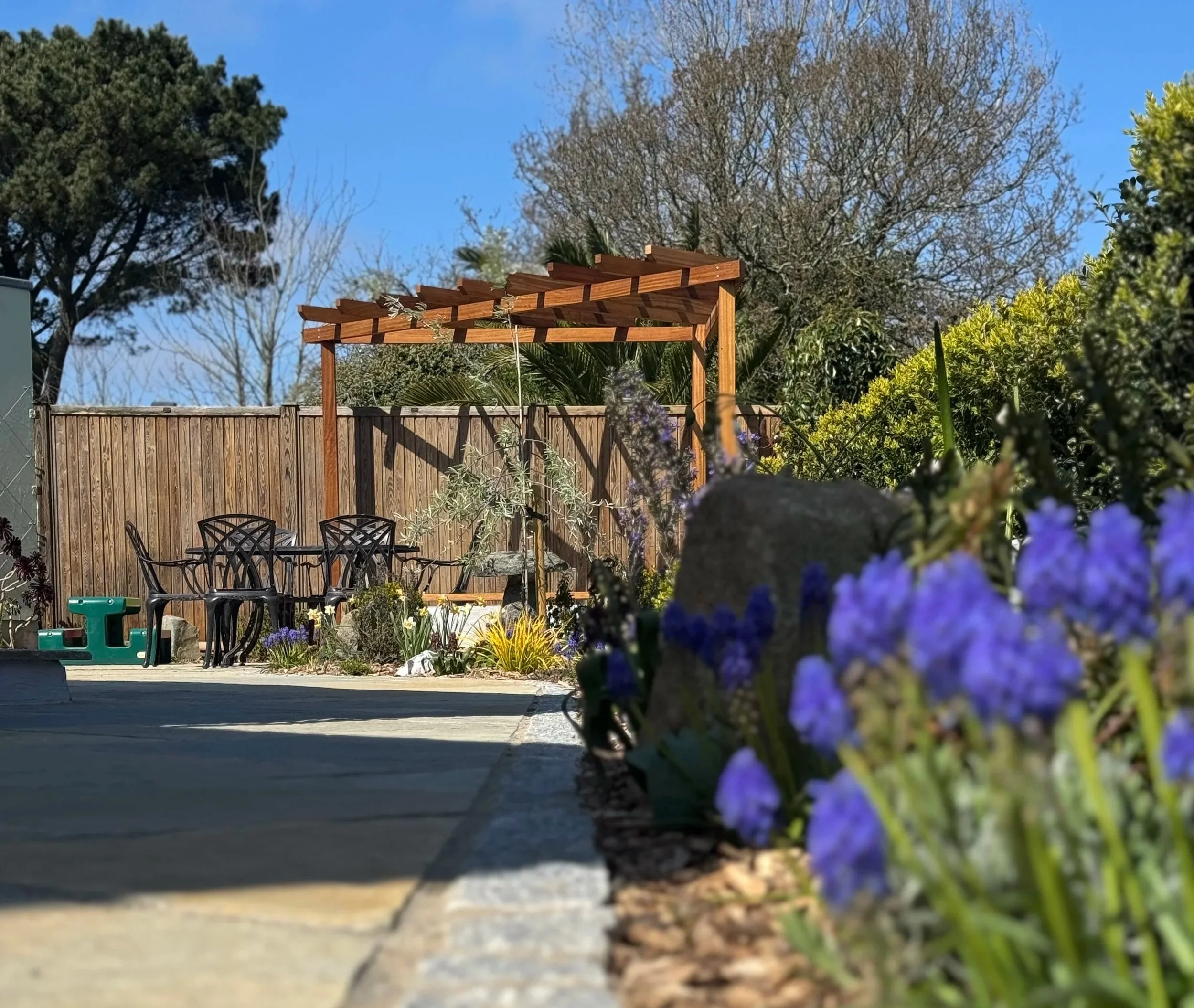 Garden with wooden pergola, patio table with chairs, colorful flowers, and clear blue sky.