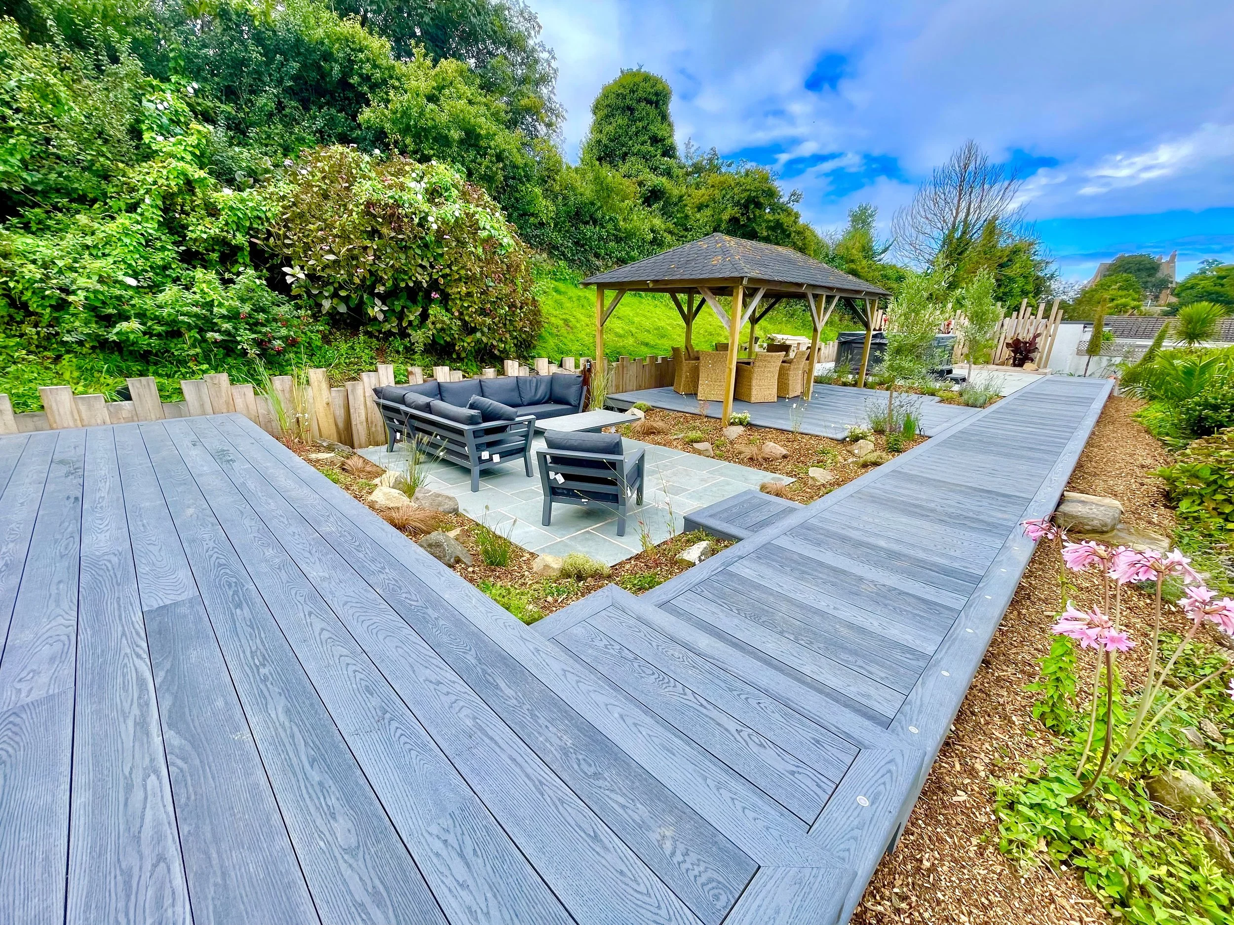 Garden with Millboard deck, seating area, and gazebo with outdoor dining table and chairs, lush green plants and trees, cloudy sky.