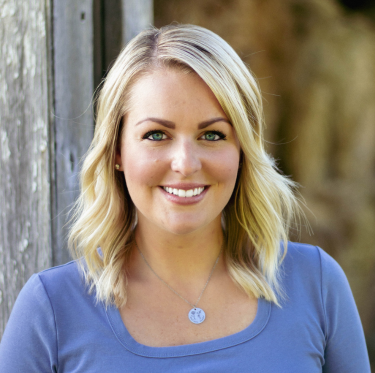 A smiling woman with blonde hair, wearing a blue shirt and a necklace, standing outdoors near a wooden fence.