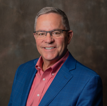 A middle-aged man with gray hair and glasses wearing a blue suit jacket and a pink shirt, smiling against a textured brown background.