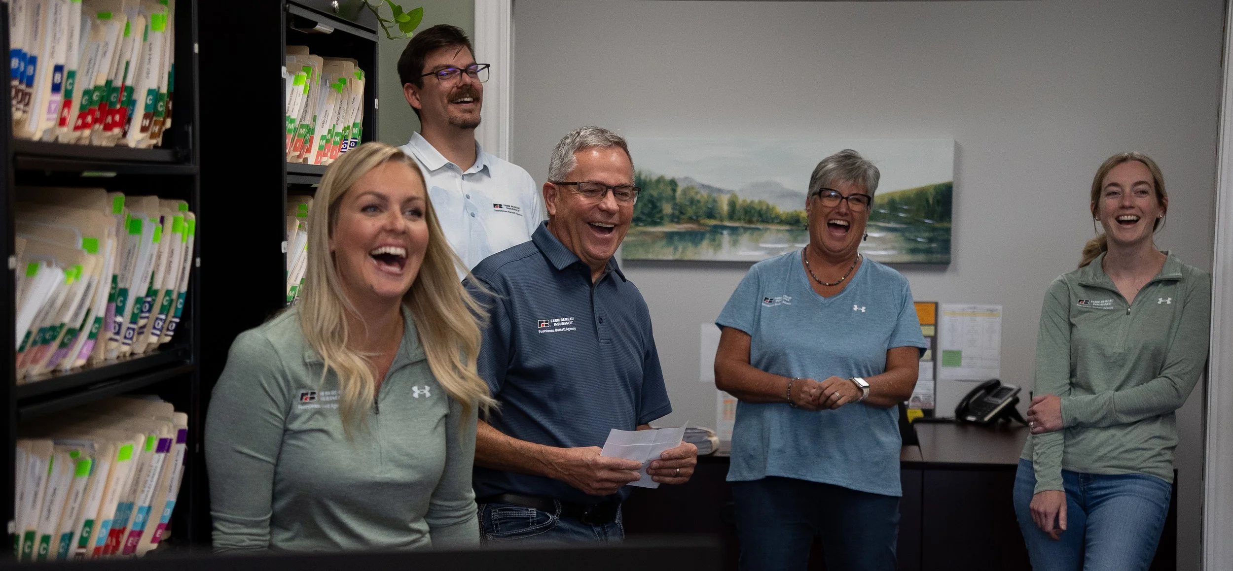 Five people in an office, smiling and laughing, with shelves of files and a landscape painting in the background.
