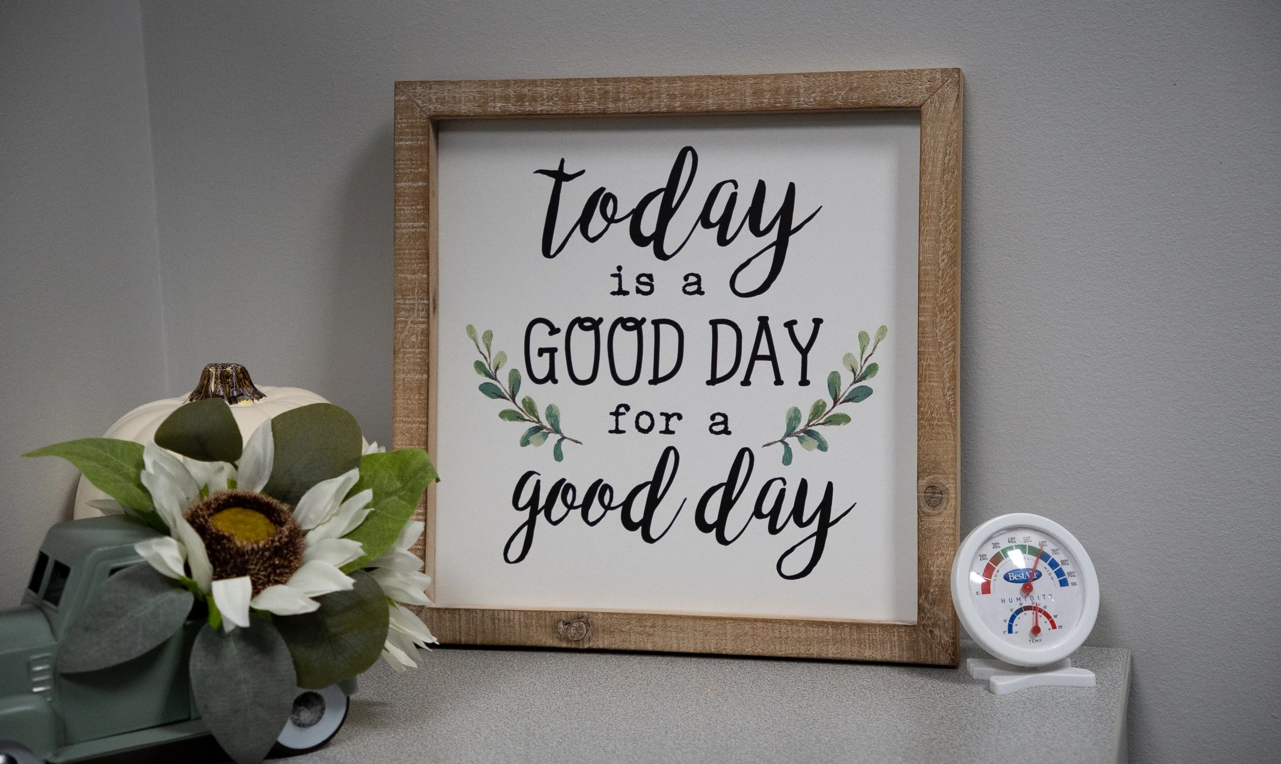 Decorative sign with the quote 'today is a good day for a good day,' framed in wood, placed on a wall behind a small table with an artificial sunflower and leaves in a vintage-style toy truck, and a white thermometer/hygrometer measuring temperature and humidity on the right.