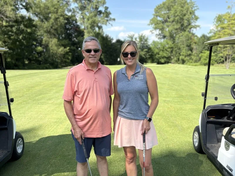 A man and a woman standing on a golf course, both holding golf clubs, with golf carts beside them and trees in the background.