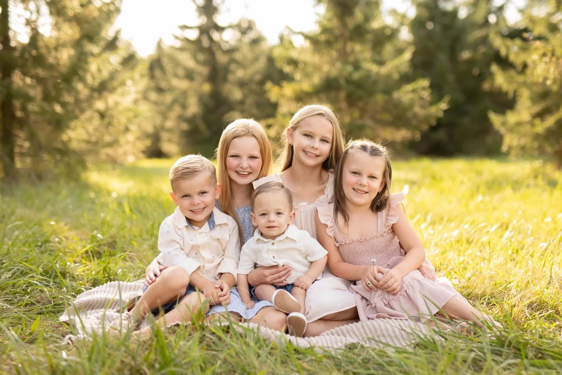 A group of five children, sitting on a blanket in a grassy field with trees in the background, smiling at the camera on a sunny day.