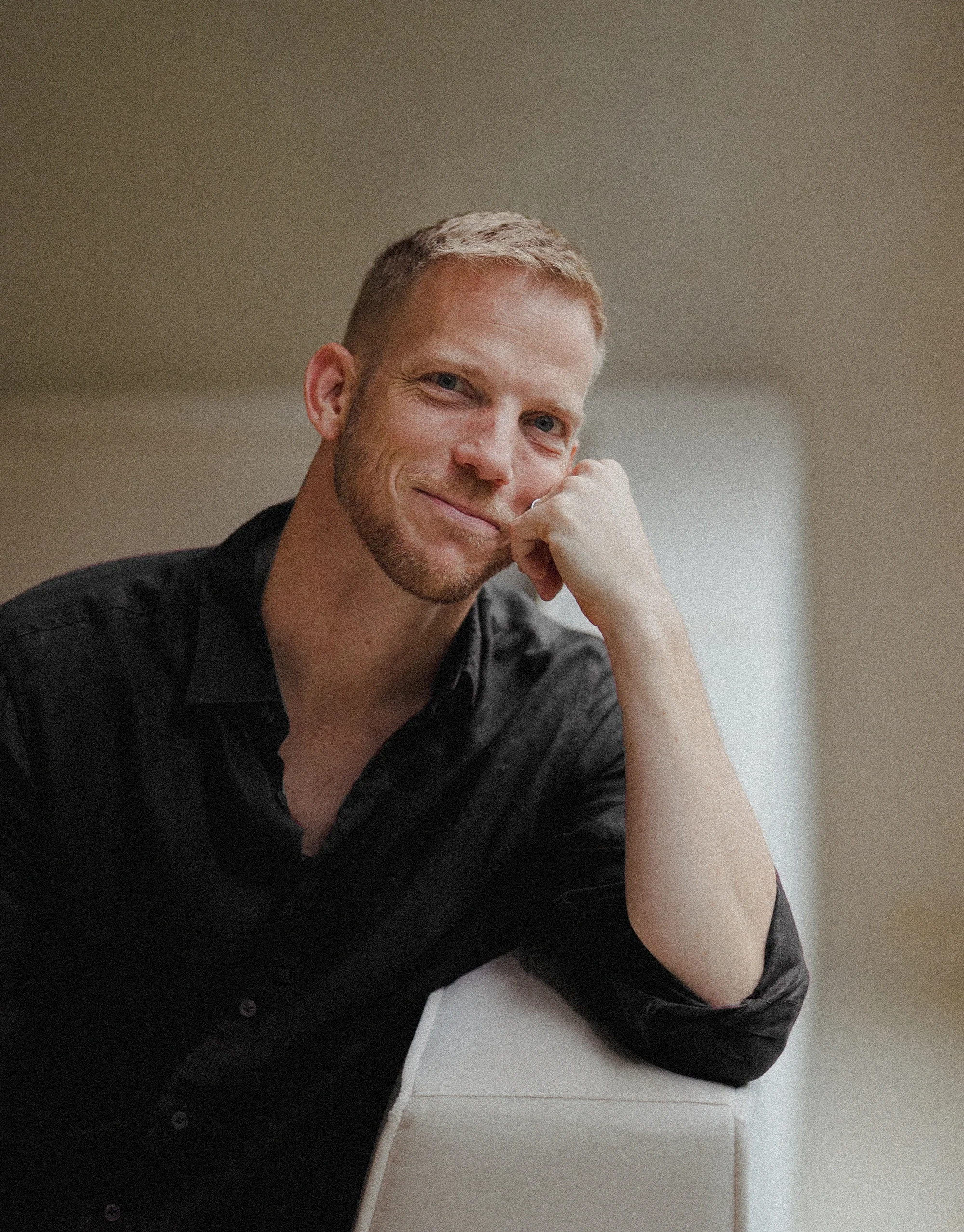A man with short, light-colored hair and a beard, wearing a black shirt, sitting with his elbow on a white surface and his head resting on his hand, smiling softly at the camera.