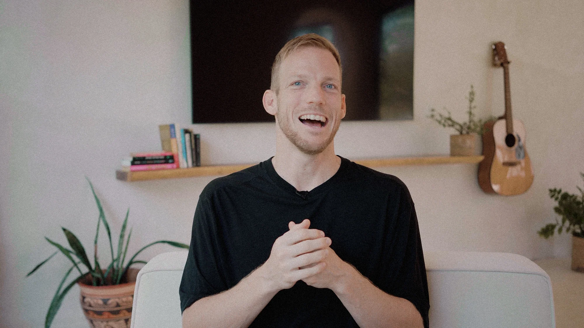 A smiling man with light hair and a beard, wearing a black shirt, sitting in a living room with a guitar hanging on the wall and a TV behind him.