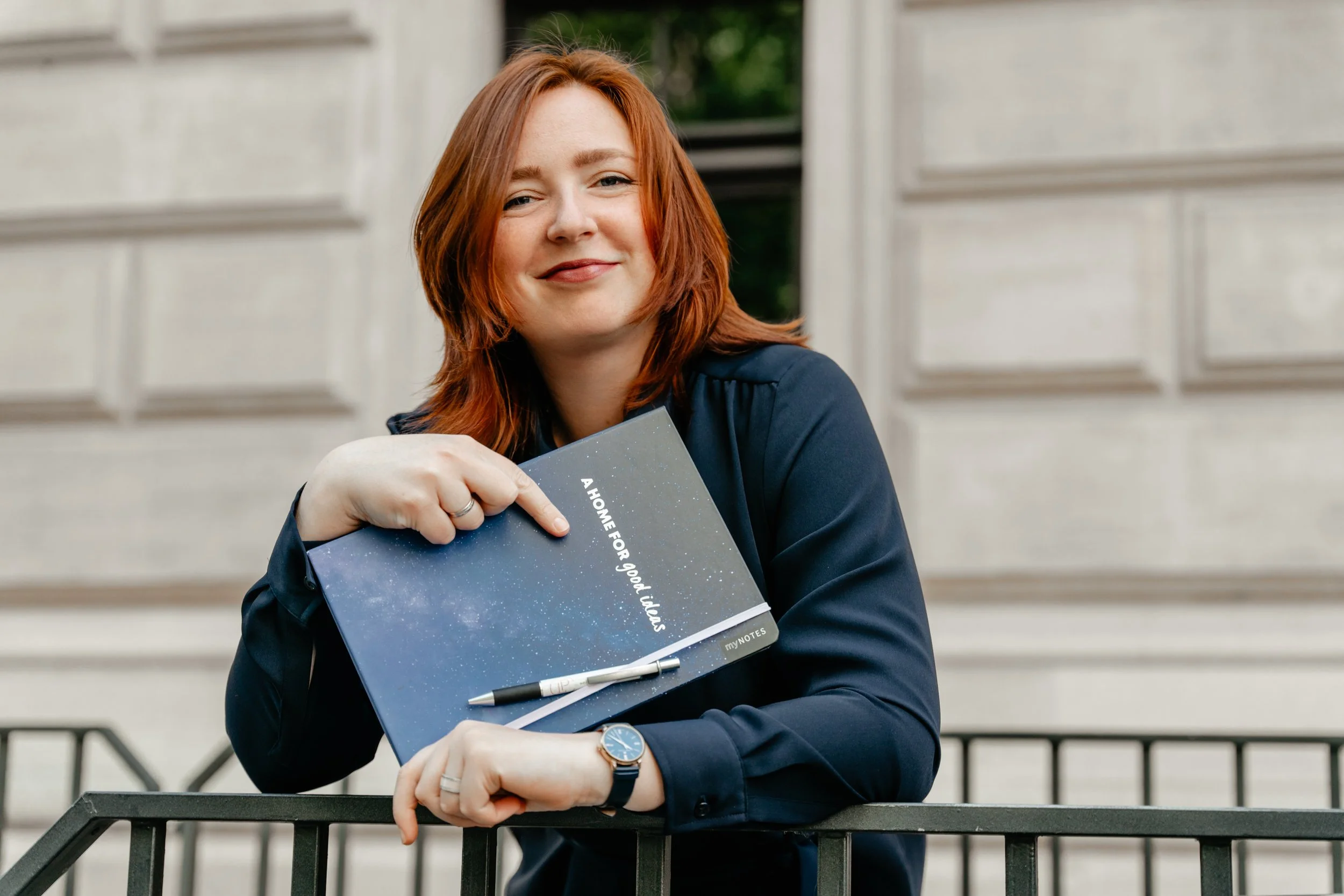 A woman with red hair smiling and holding a notebook that says, 'A home for good ideas,' standing outdoors in front of a building, leaning on a black metal railing.