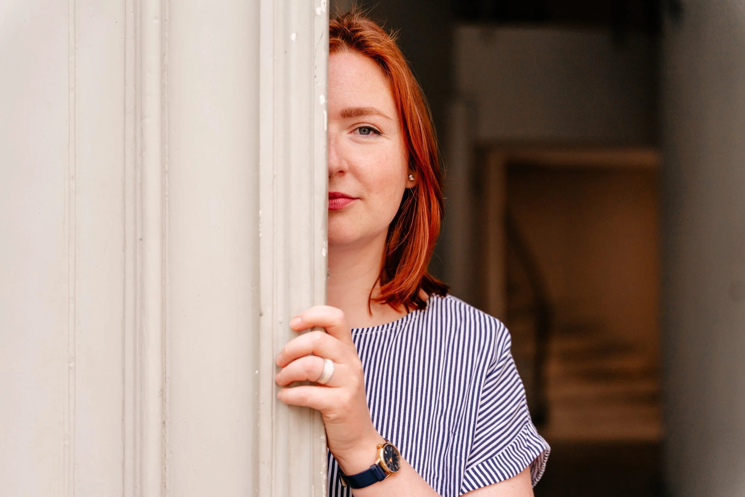 A woman with red hair partially hides behind a white door, peering out with a slight smile, wearing a striped shirt and a watch.