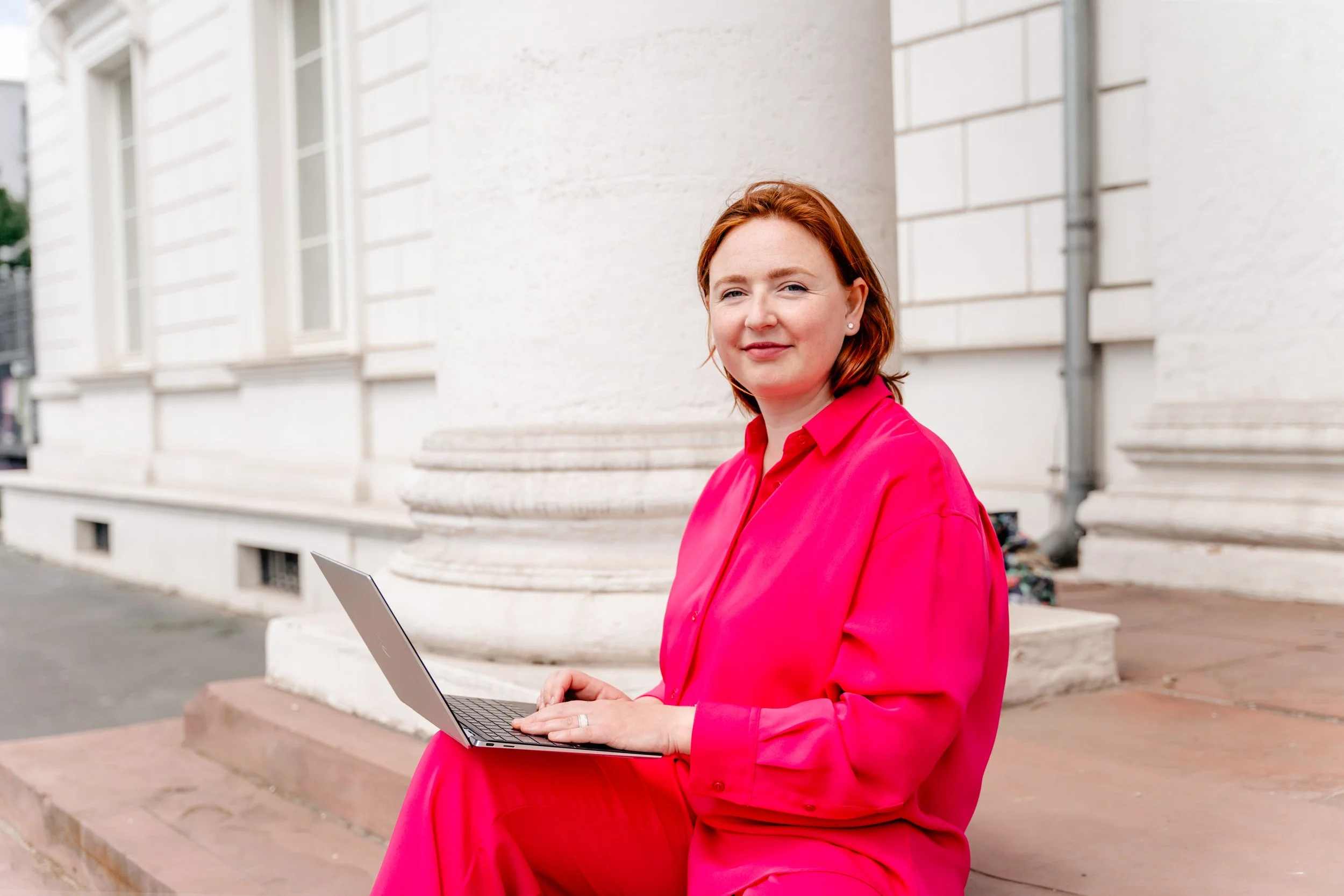 A woman with red hair in a bright pink outfit sitting outside on the steps with a laptop.