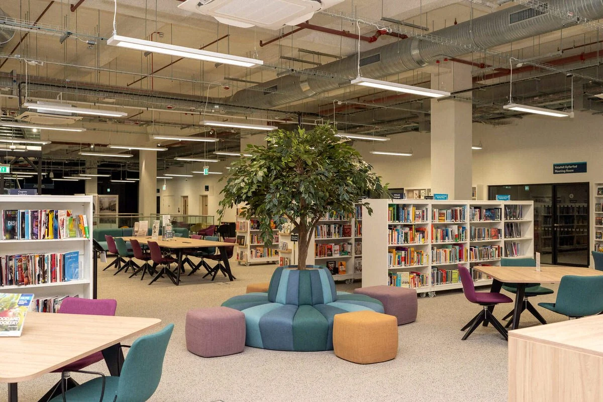 Interior view of Y Storfa main library, bookcases full of books, a circular seating area with a fake tree, colourful seating and exposed lighting. Pictures taken to show the furniture provided by Morleys.