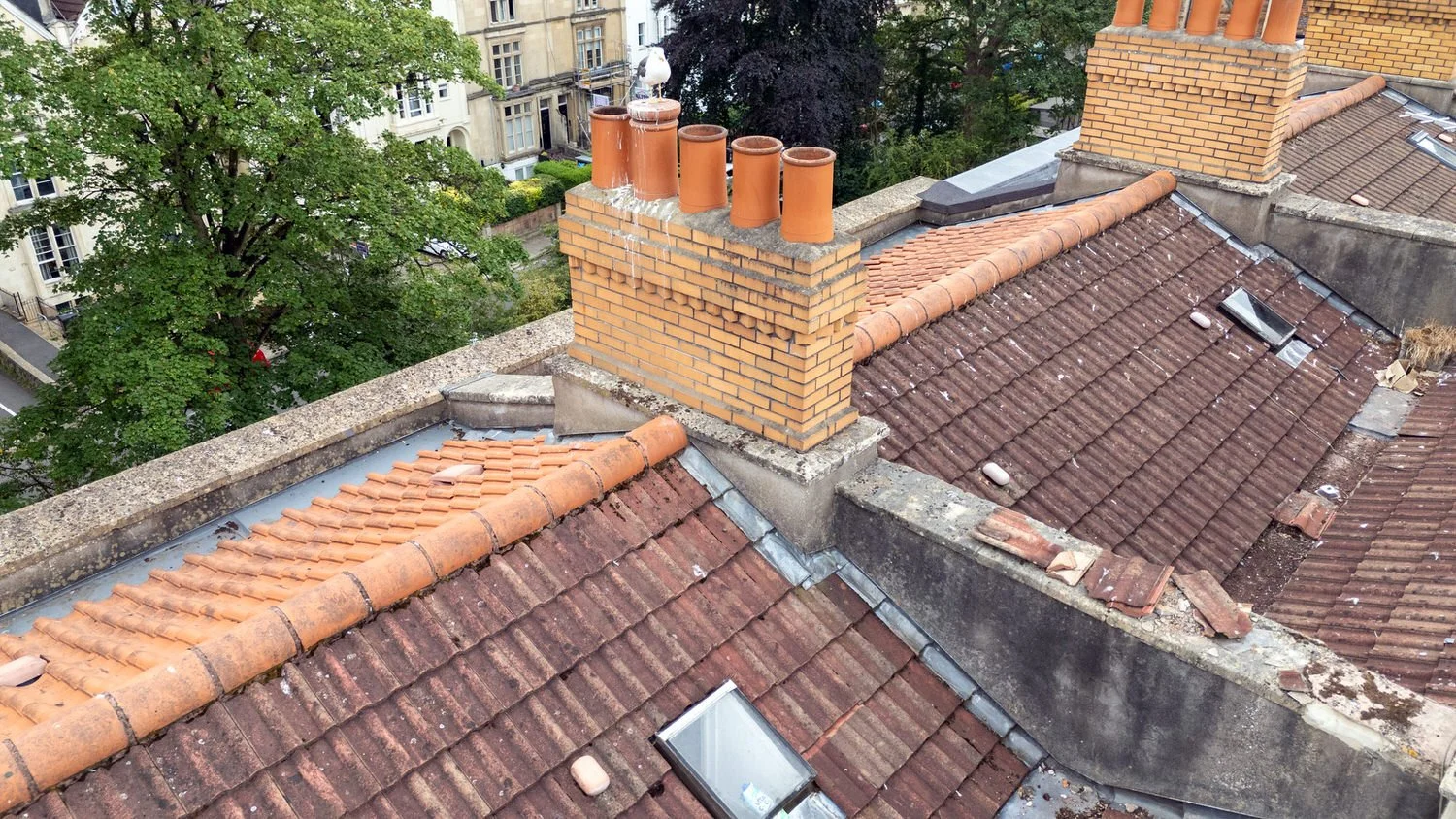 A drone survey roof view reveals red tile rooftops and yellow brick chimneys, nestled among trees in an urban residential area.