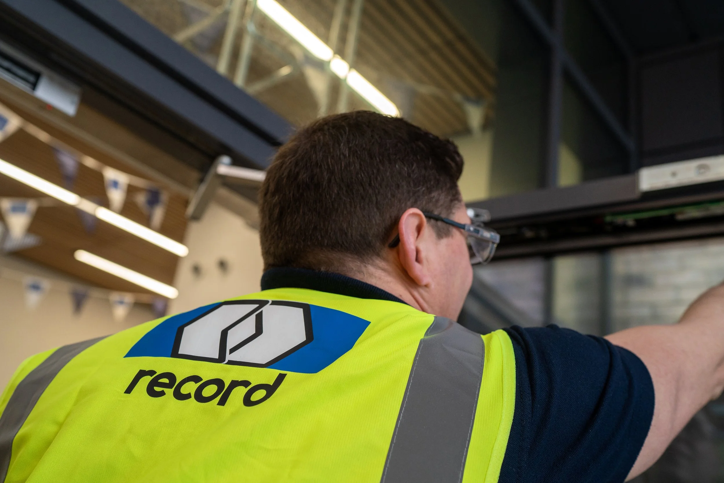 A person in a yellow safety vest with record logo works on a glass door indoors.