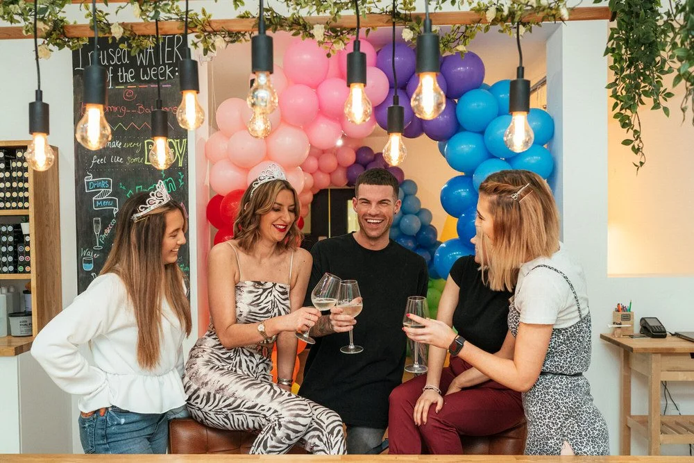 Five colleagues celebrating with wine glasses in a decorated party space with colorful balloons and hanging lights.