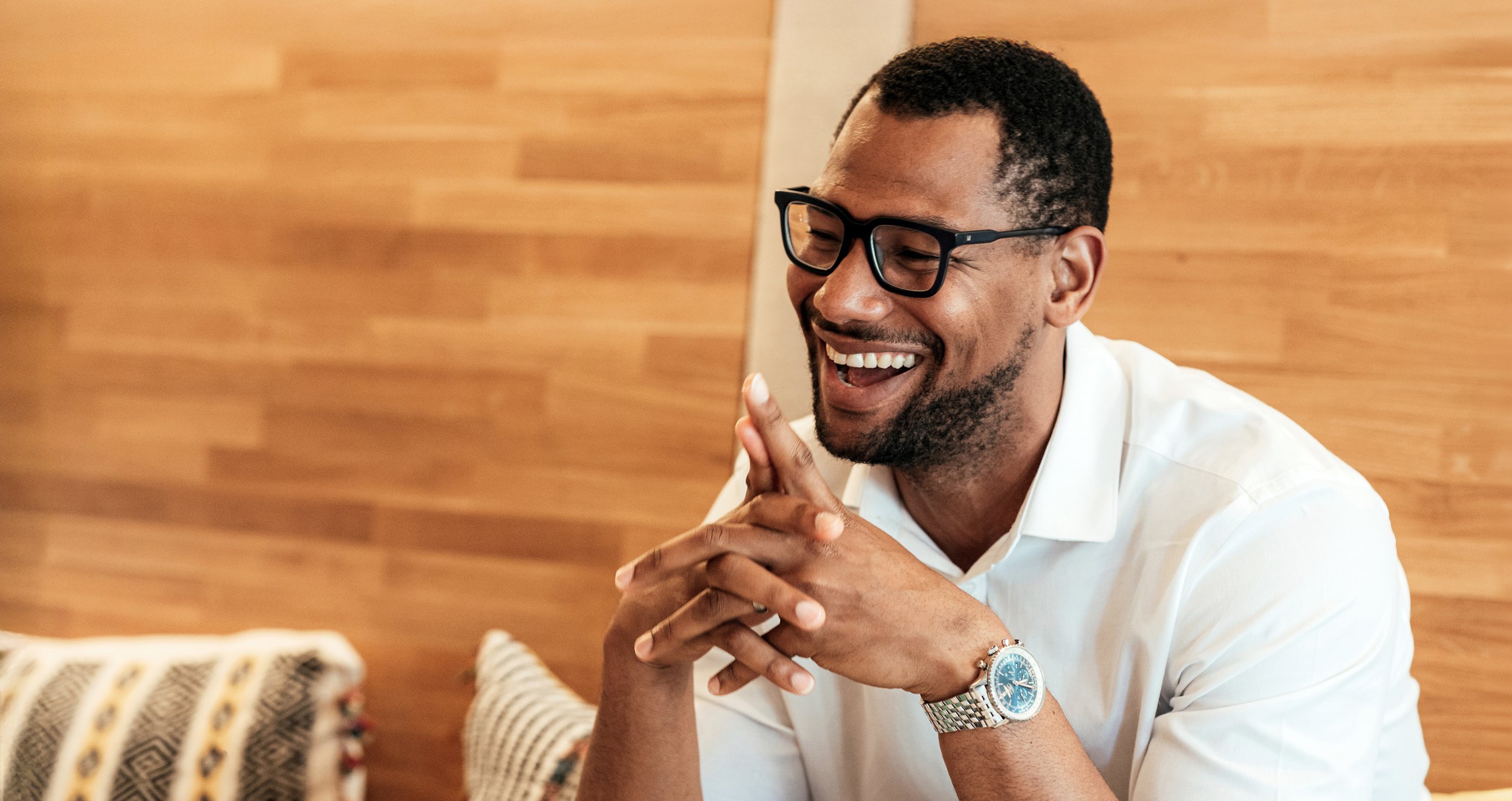 A man wearing glasses and a white shirt, smiling and laughing with his hands clasped together, sitting indoors against a wooden wall.