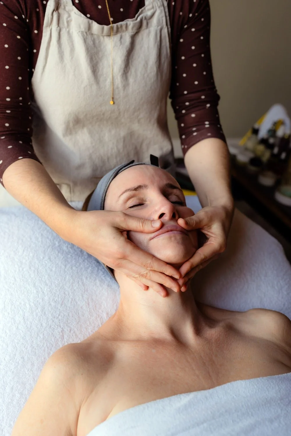 Massage professional's hands providing a facial to a woman laying on a bed in a white towel.