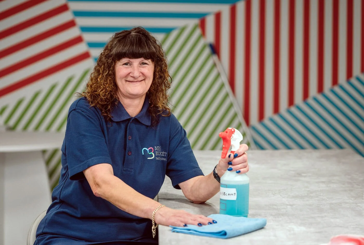 A woman in a blue polo shirt smiles at the camera, cleaning a table—a friendly business headshot in action.