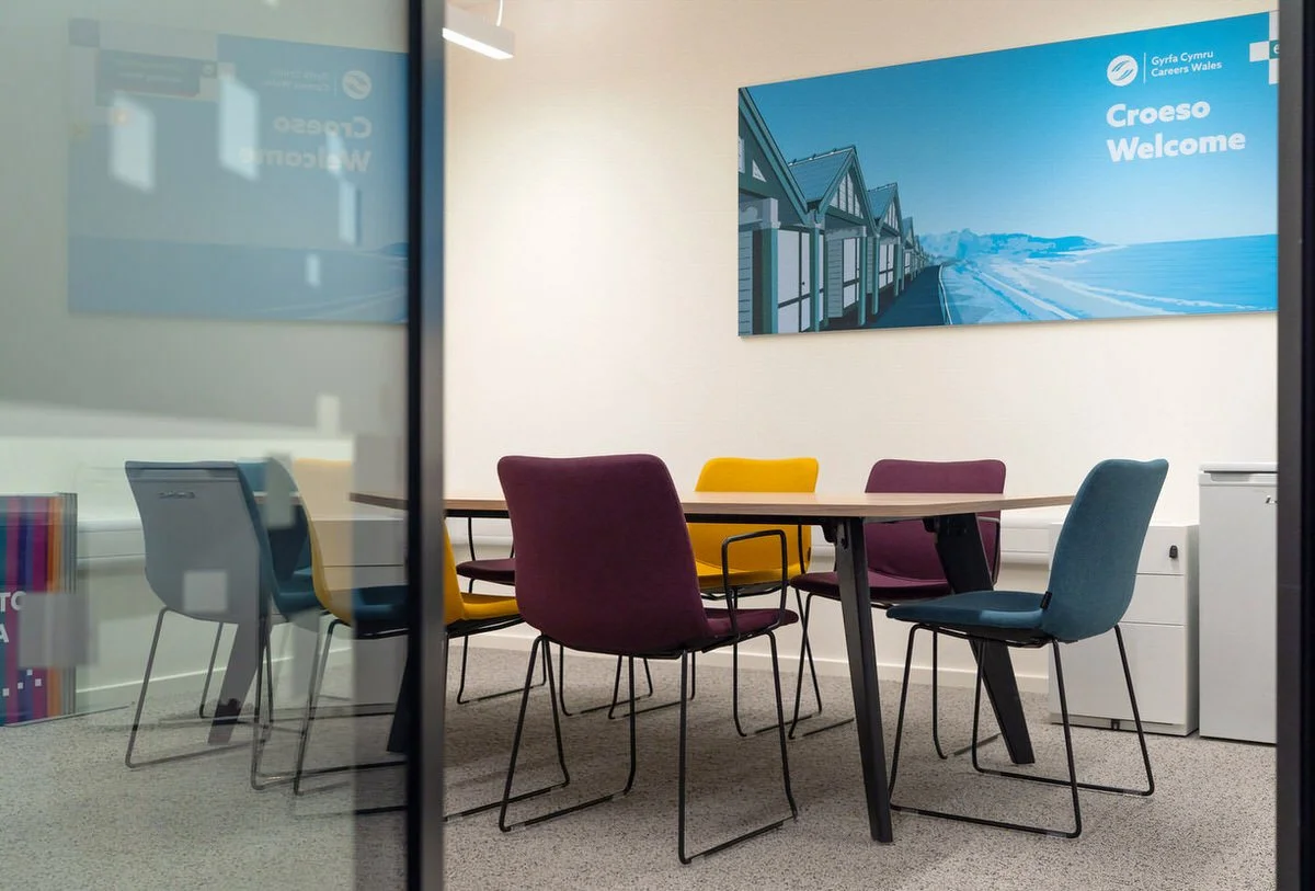 Office space inside Y Storfa hub in Swansea. Shown through a glass door, the table with 6 colourful chairs in a private space featuring artwork on the wall saying Croeso, Welcome.