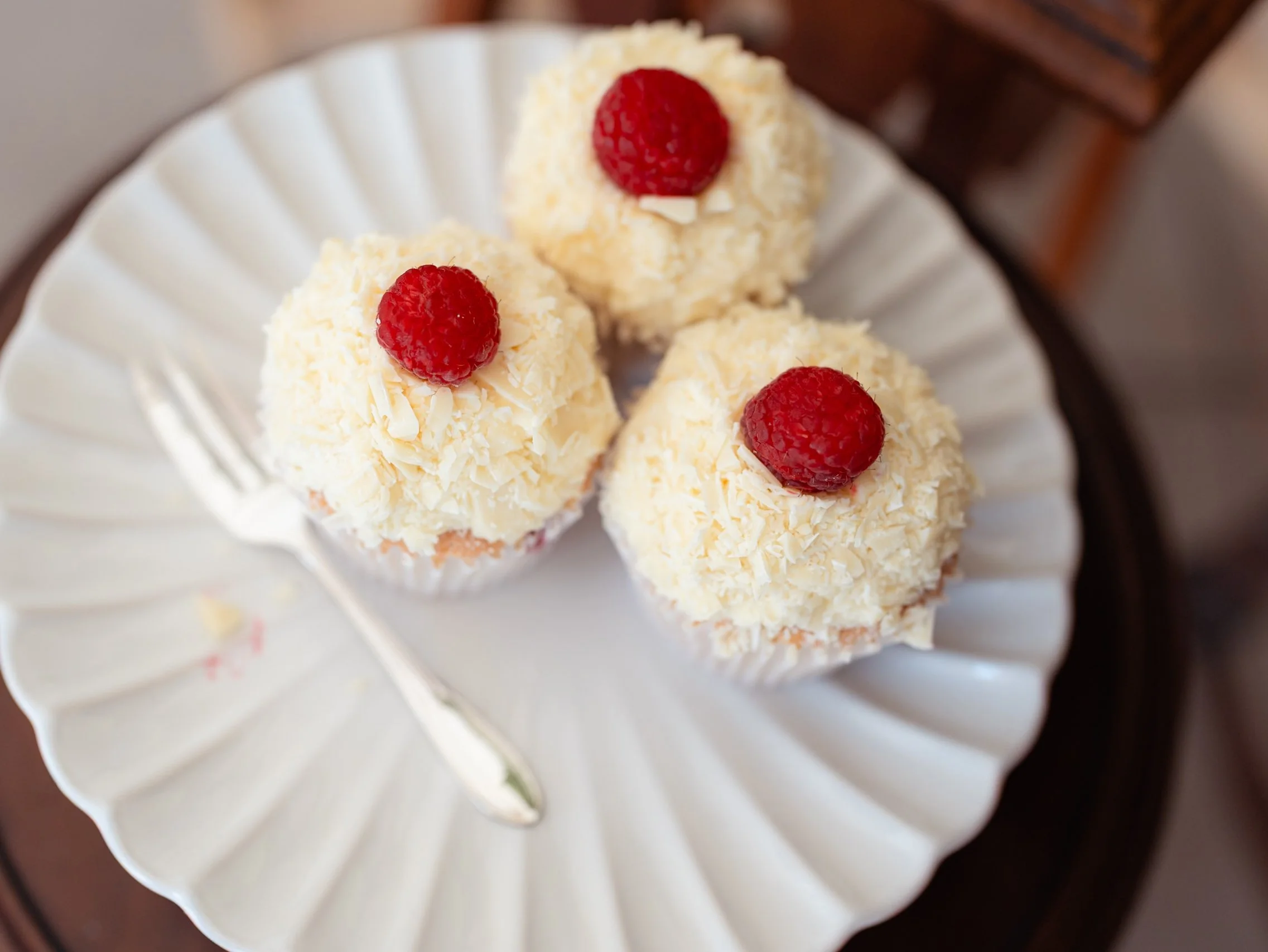 Baker's Product photography of three cupcakes topped with white chocolate shavings and a raspberry, served on a white plate with a fork.