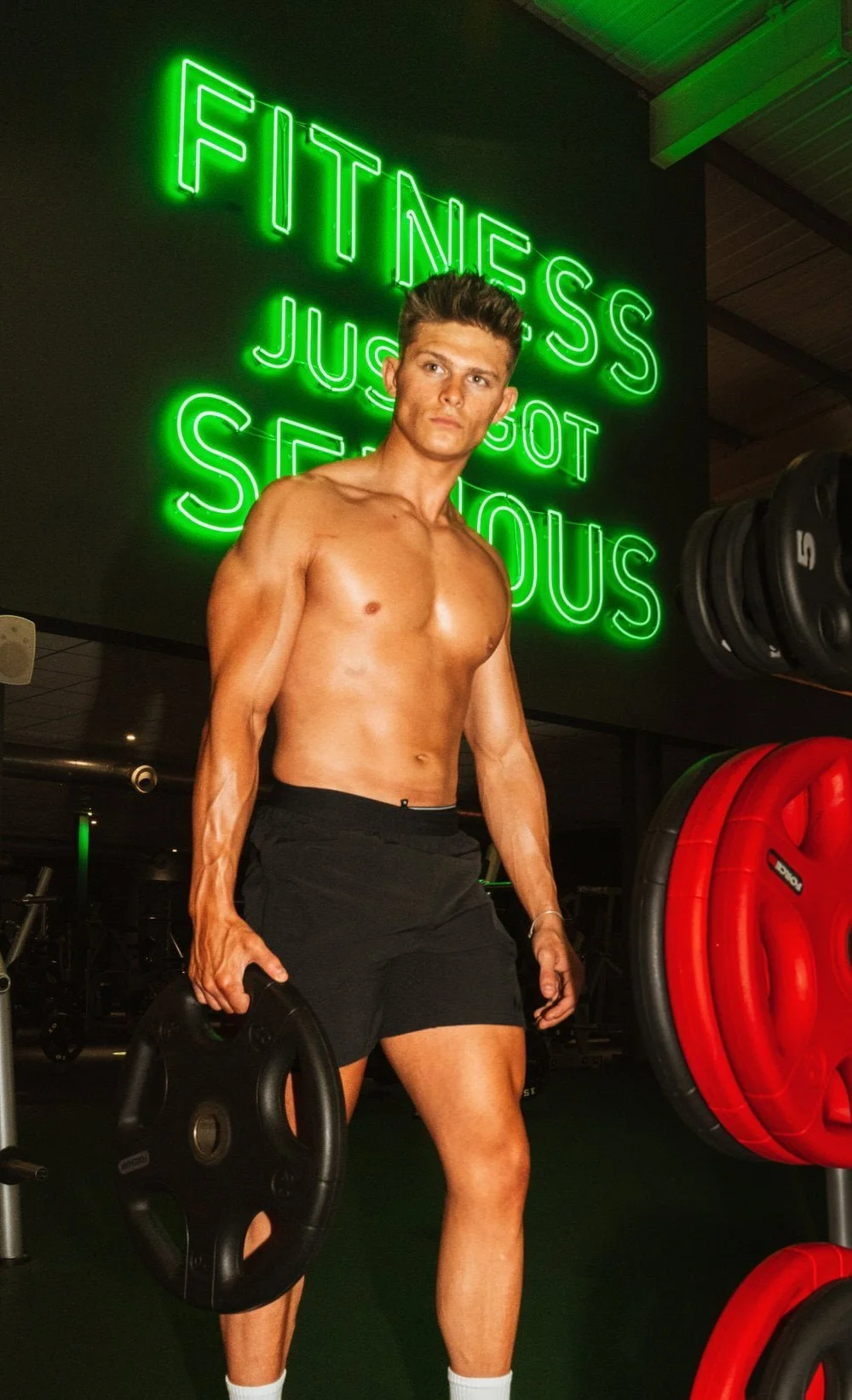 Bare chested man in a gym holding a weight, with a neon sign behind reading Fitness just got serious, in a JD gym in Swansea.