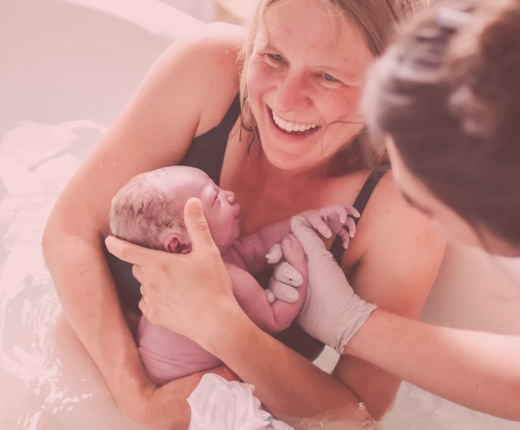 Eine Frau hält ein neugeborenes Baby in den Armen, während eine Hebamme sie begleitet. Die Szene findet wahrscheinlich während einer Geburt in einem Wassergeburtsbereich statt.