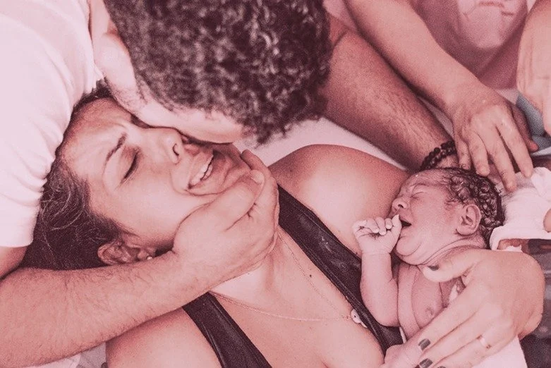 Eine Frau liegt mit einem Neugeborenen auf dem Arm, während ein Mann sie küsst. Das Neugeborene schreit und hält die Hand zur Faust. Alle scheinen emotional berührt.