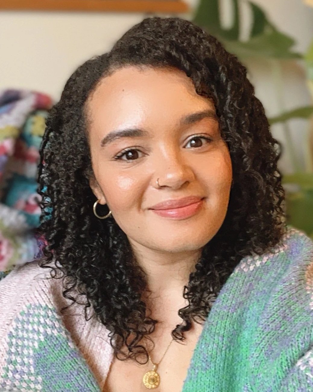 A woman with dark curly hair, light brown skin, and wearing hoop earrings and a gold pendant necklace, smiling at the camera in an indoor setting.