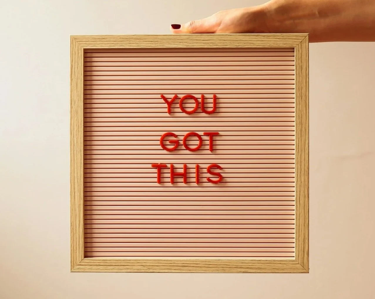 A framed letter board with pink background and red plastic letters spelling "YOU GOT THIS." A hand with dark nail polish is holding the top of the frame.