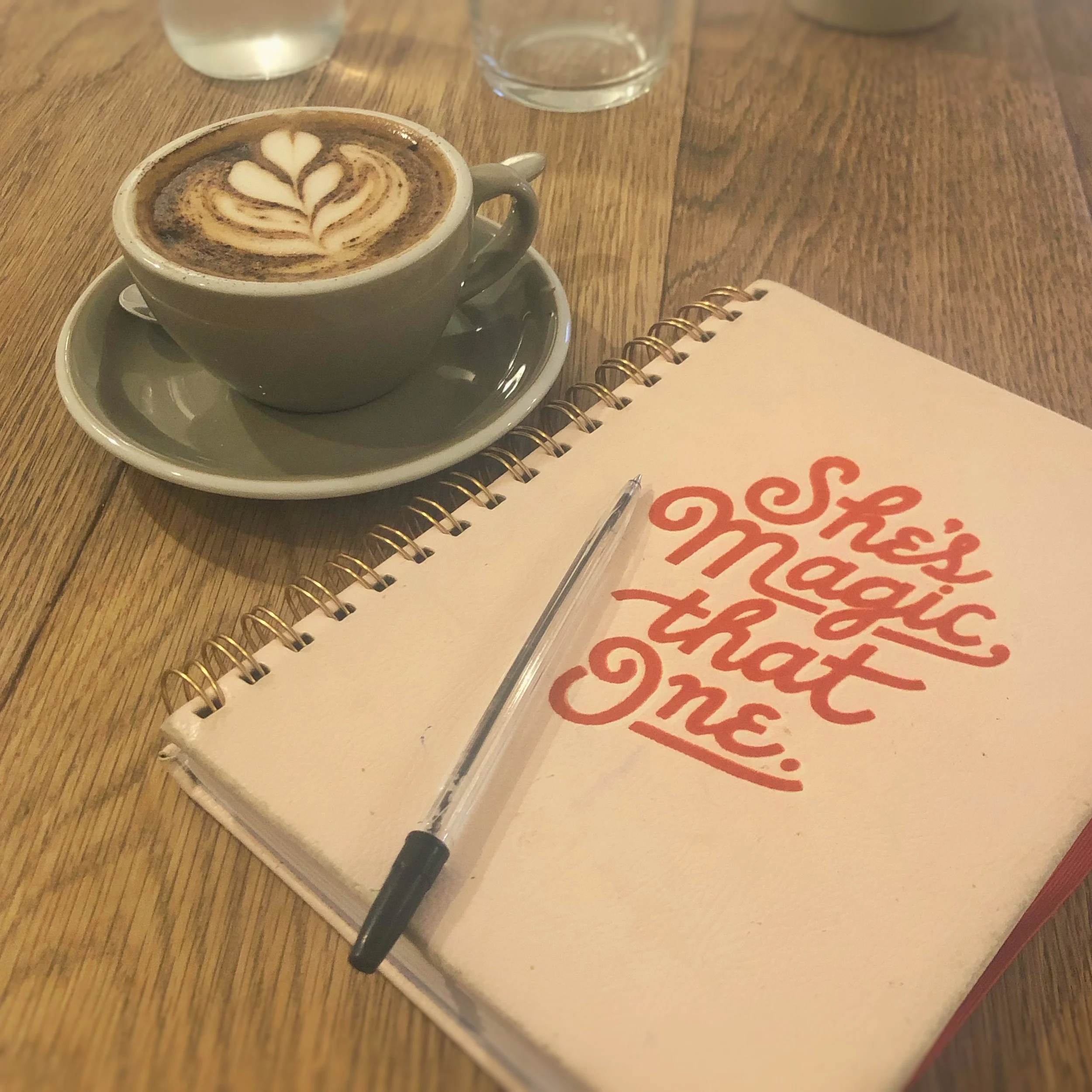 A cup of coffee with latte art on a saucer, a glass of water, a spiral notebook with 'She's magic that one' written in red, and a pen on a wooden table.