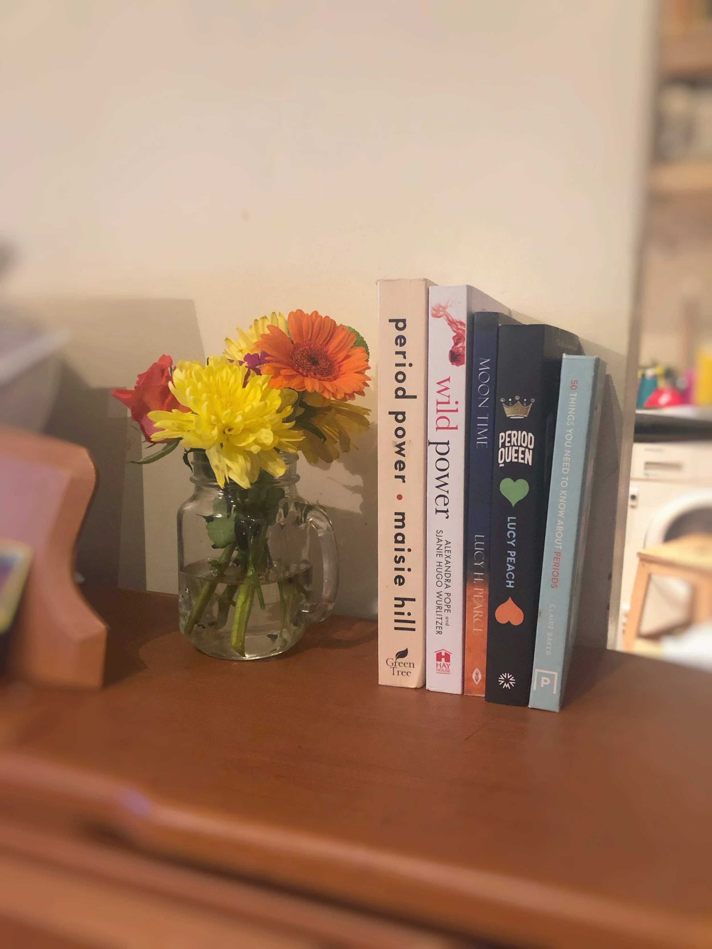 A clear glass mason jar with water, holding a bouquet of colorful flowers including yellow, orange, and red blooms, placed on a wooden surface next to a small stack of five books about periods and menstrual cycle awareness.