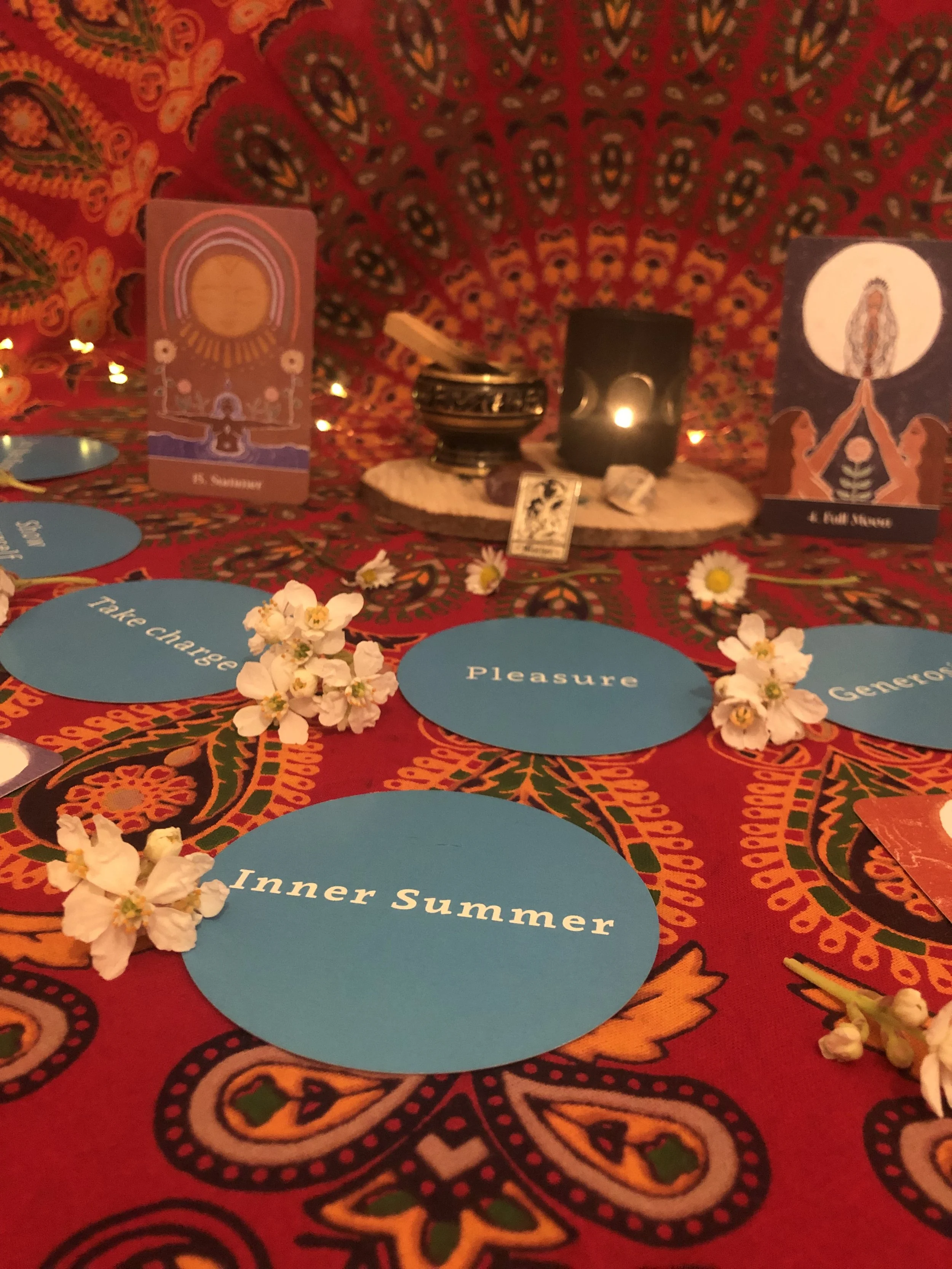 A table setup for a tarot card reading with colorful tablecloth, blue circles labeled with words like 'Inner Summer,' 'Pleasure,' and 'Give,' flower petals, and tarot cards with an altar in the background.