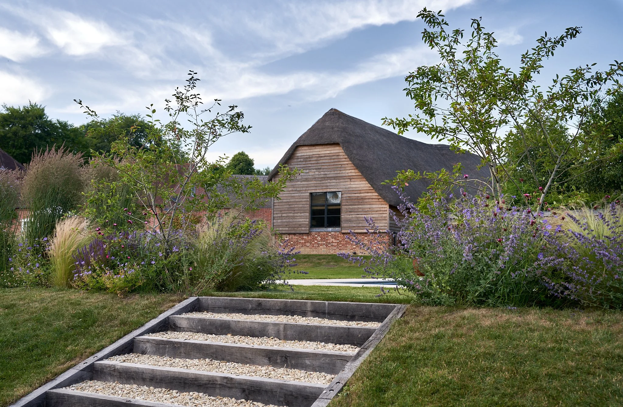 A garden pathway leading to a rustic wooden house with a thatched roof, surrounded by flowering plants and greenery, under a partly cloudy sky.