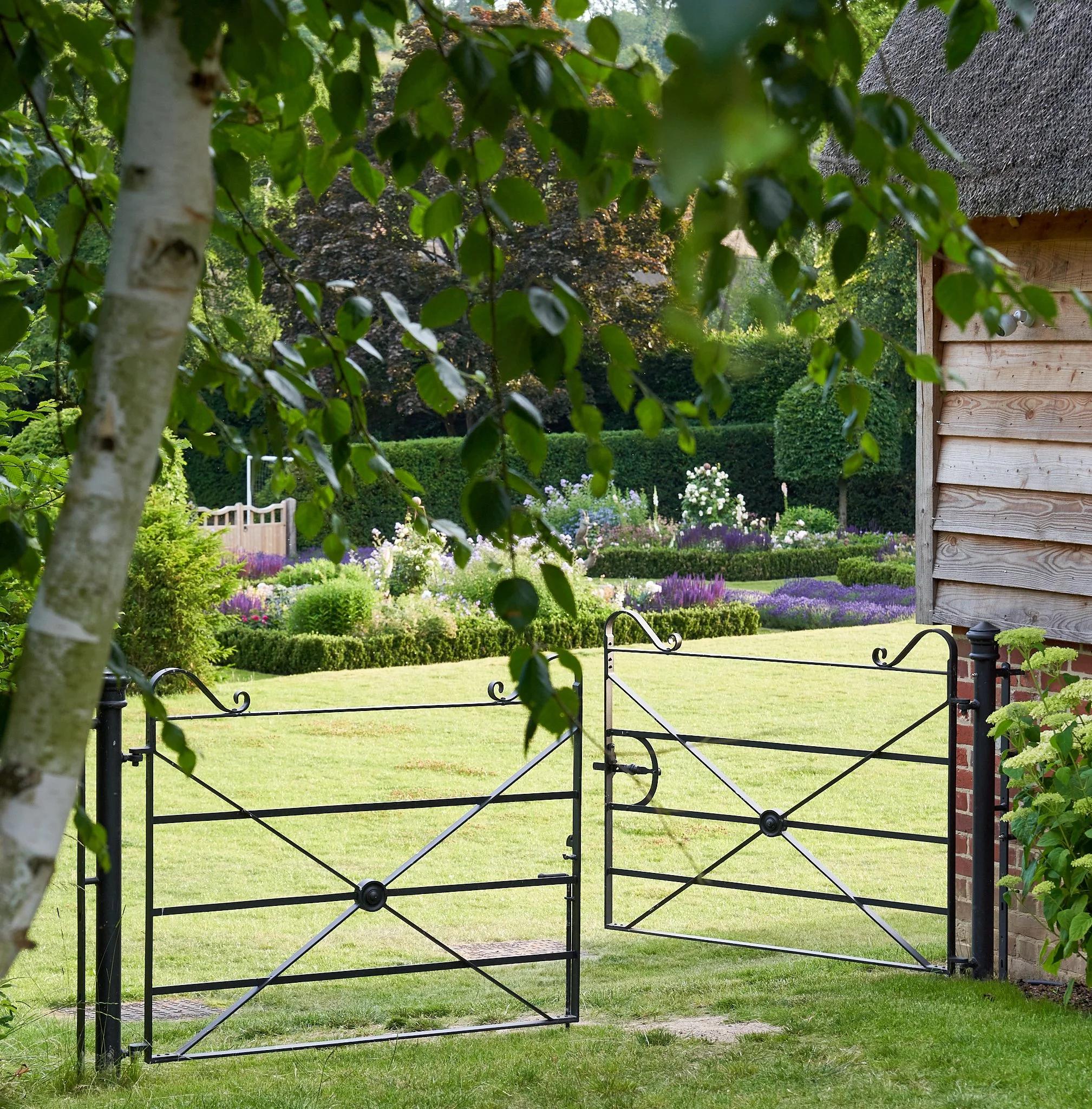 Black metal garden gate opens to a well-maintained lawn with colorful flower beds, surrounded by trees, bushes, and a wooden shed on the right.