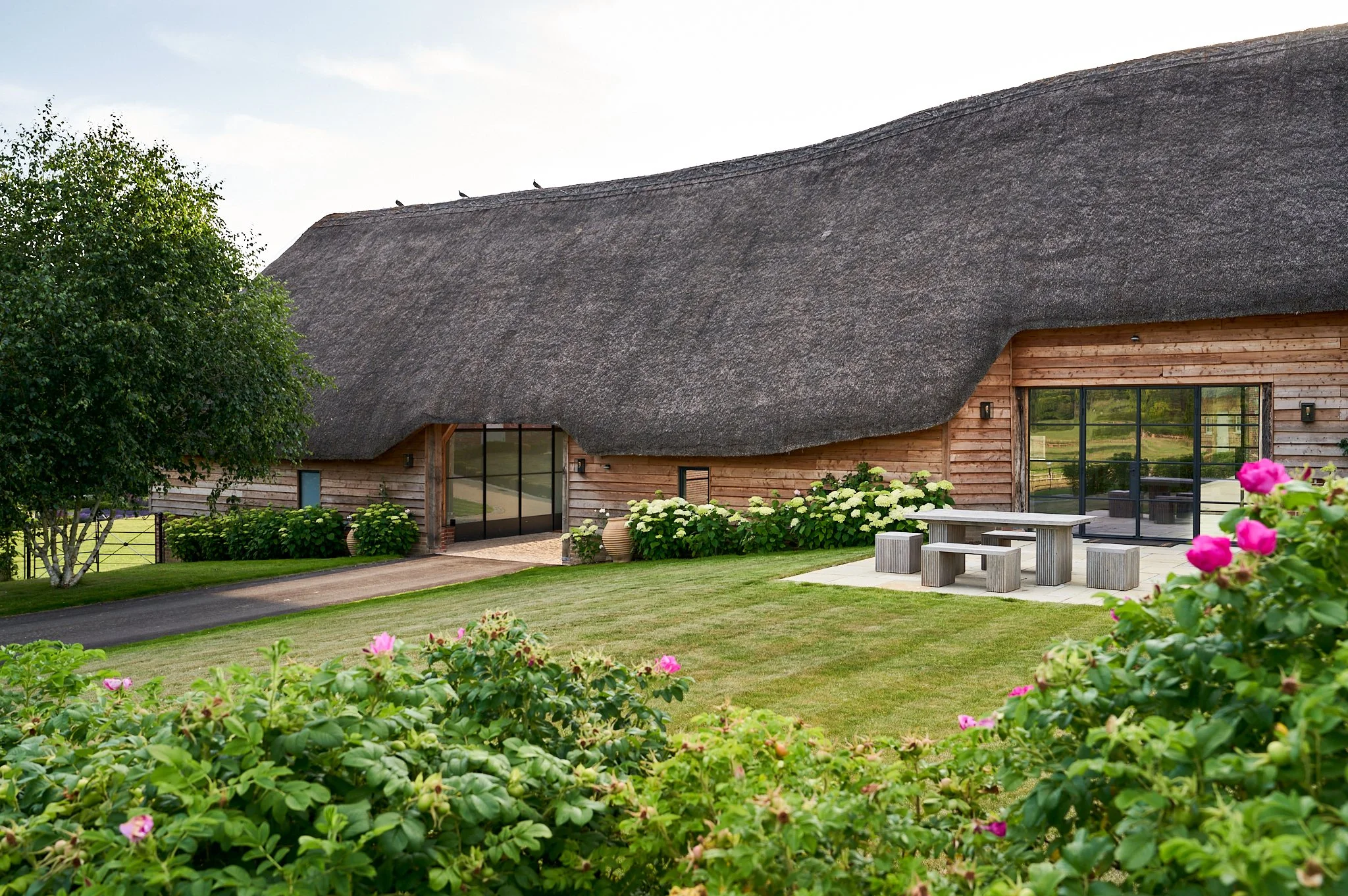 A house with a thatched roof, wooden exterior, and large glass doors, surrounded by green grass and bushes with pink flowers.