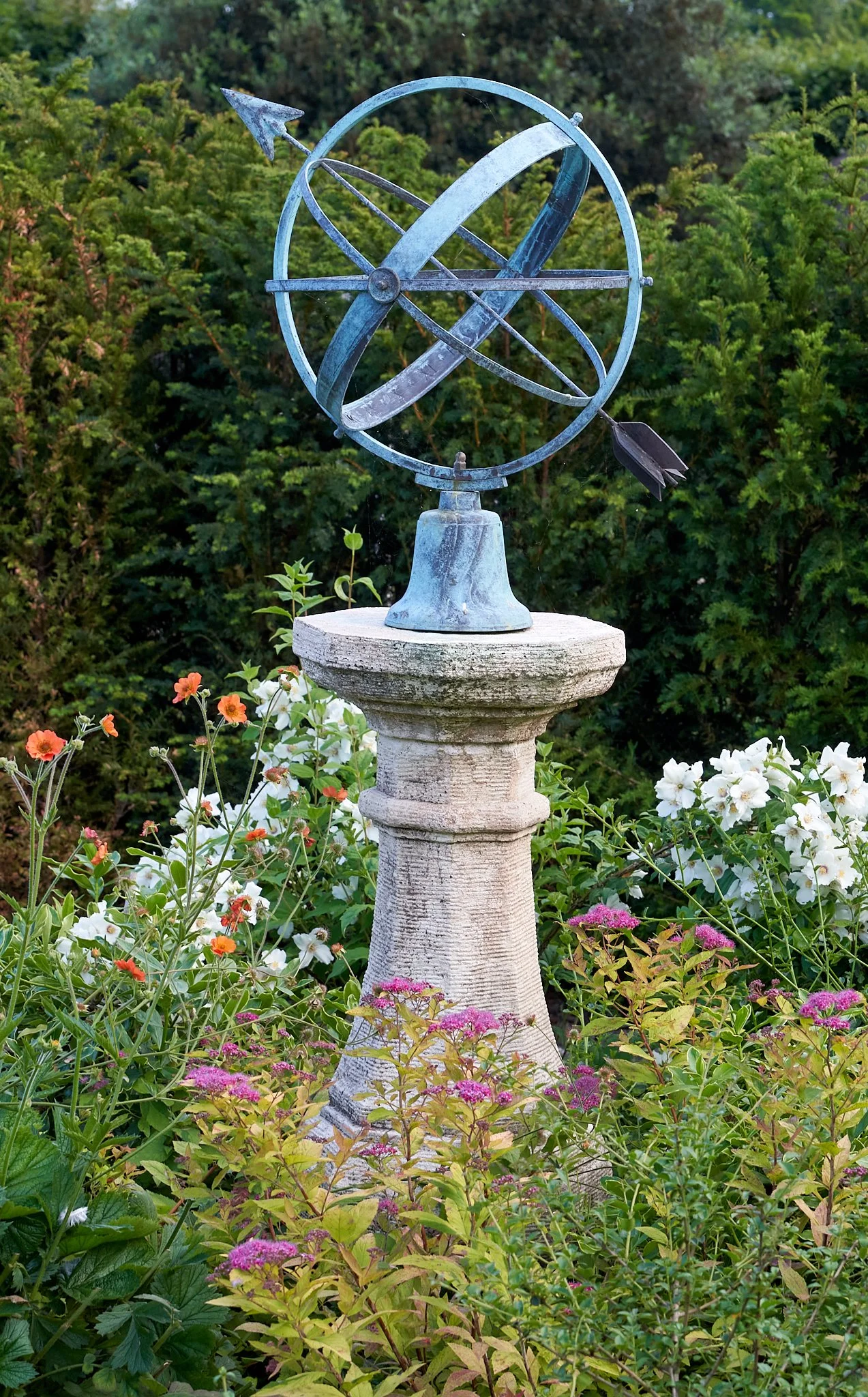A metal armillary sphere sculpture with pointed arrows, mounted on a stone pedestal, surrounded by lush green bushes and colorful flowers.