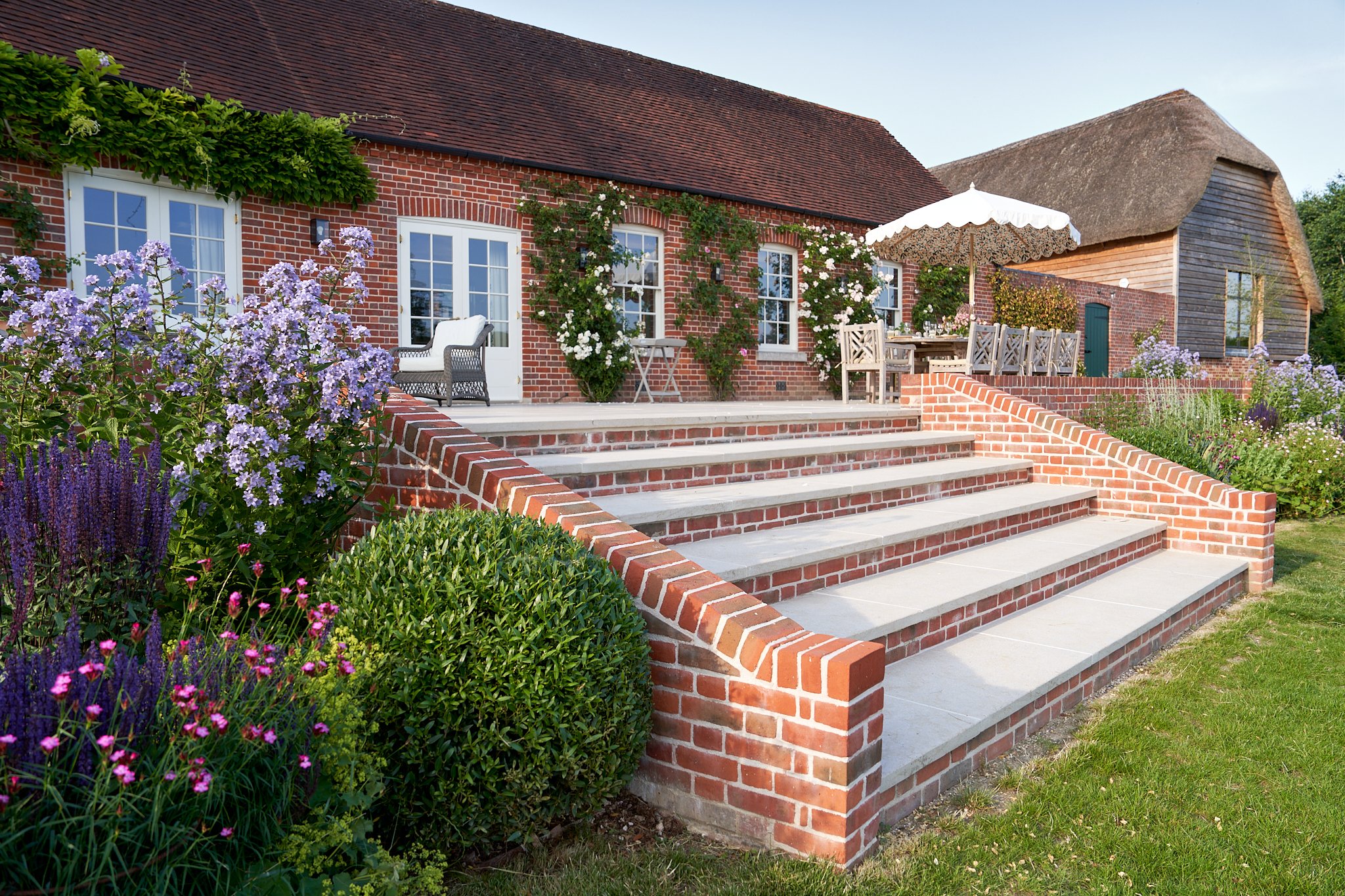 A brick house with a garden and outdoor patio with seating, flowers, and an umbrella.