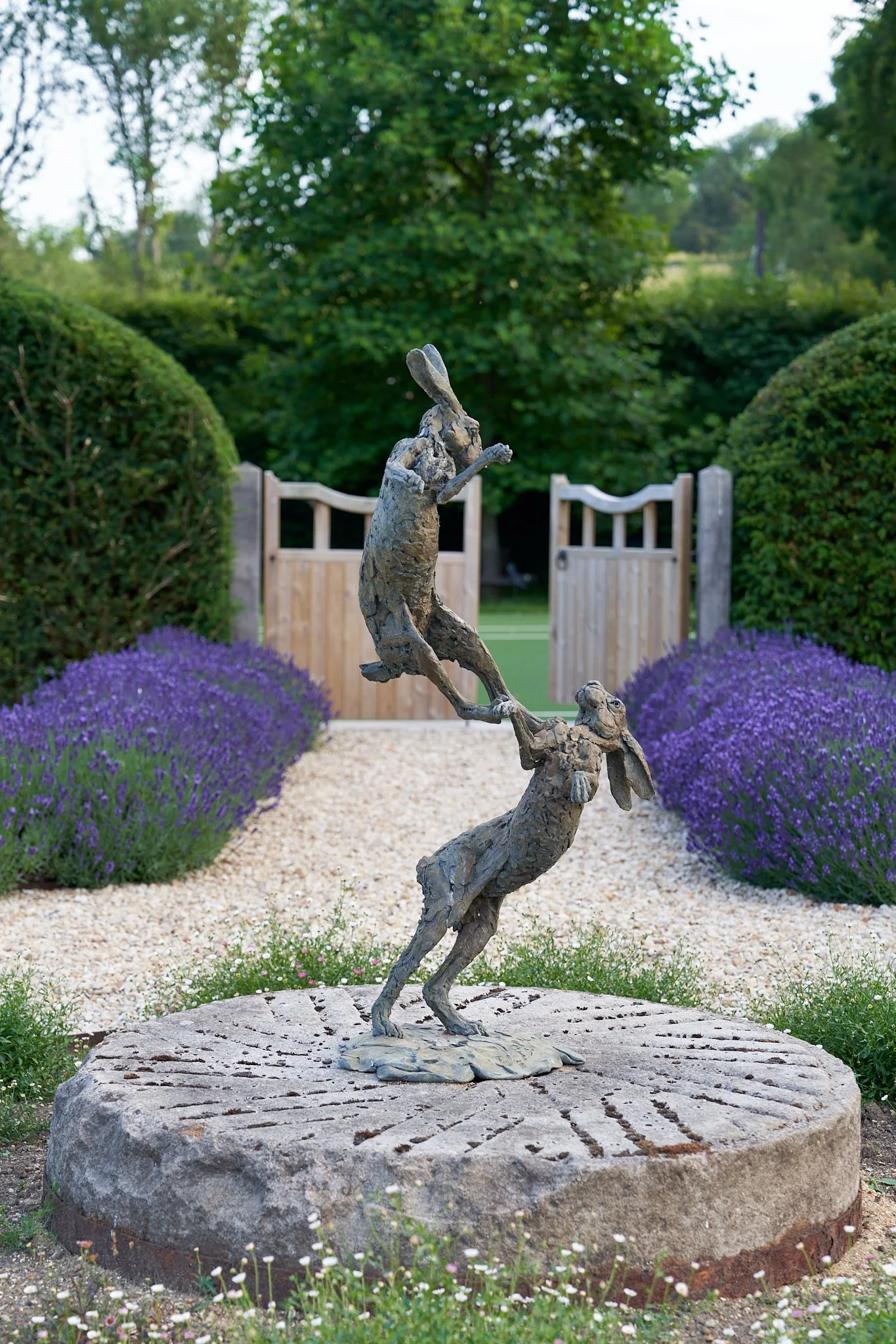 A bronze sculpture of two rabbits balancing on their hind legs in a garden, with purple flowers along a gravel path and a wooden gate in the background.