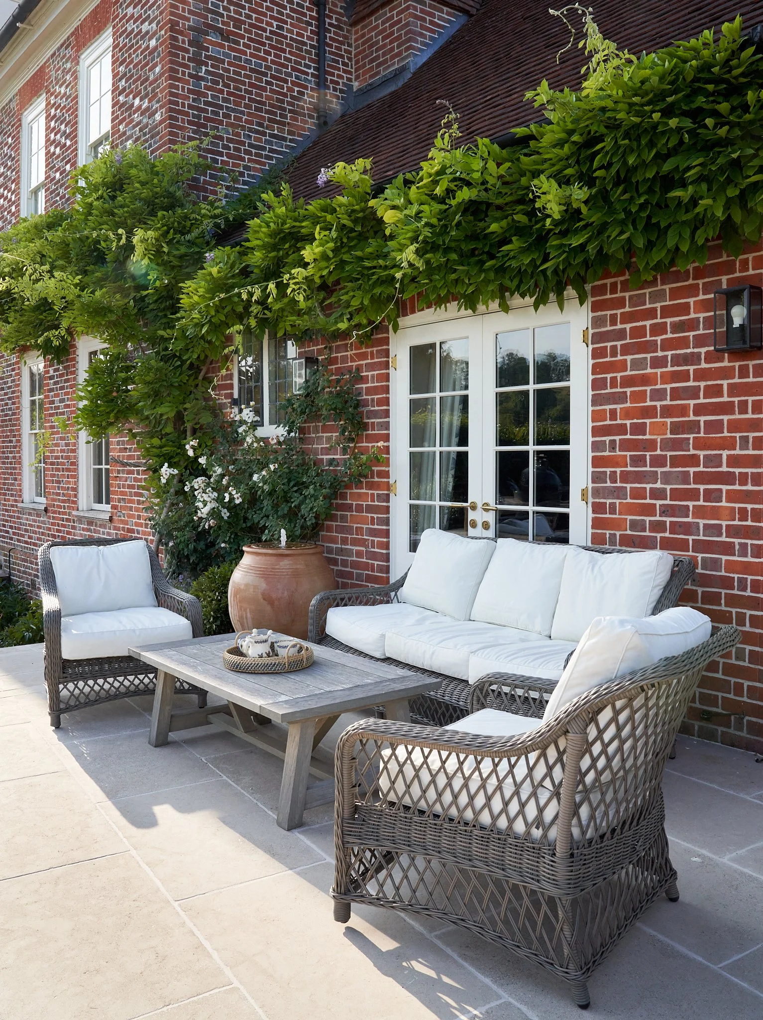 Outdoor limestone patio with wicker furniture including a sofa, armchair, and coffee table with white cushions, against a red brick Queen Anne house with white French doors, greenery, and a water feature for white noise with climbing plants.