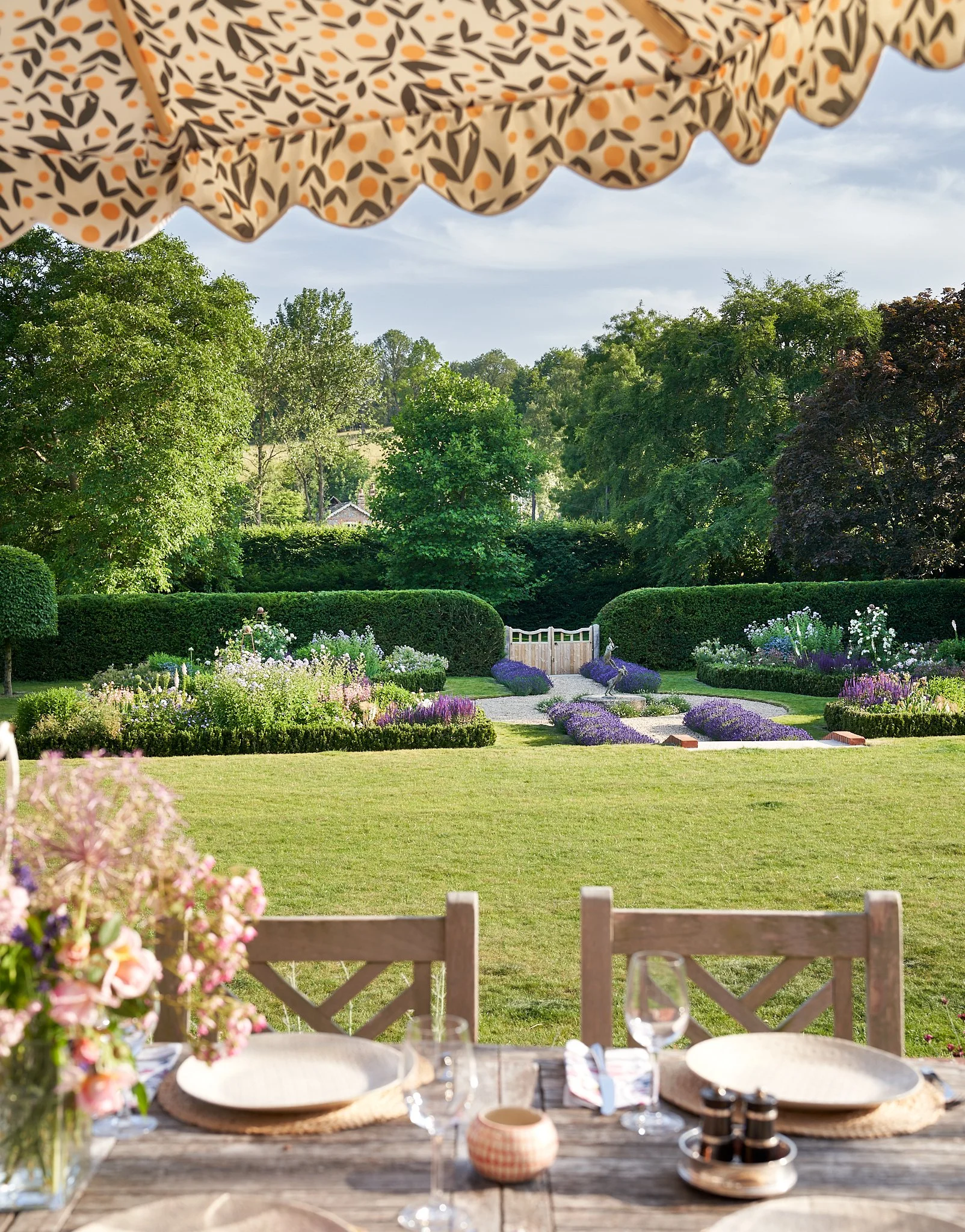 A dining table set with plates, glasses, and a flower arrangement, overlooking a well-maintained garden with flowers, shrubs, and trees under a cloudy sky.