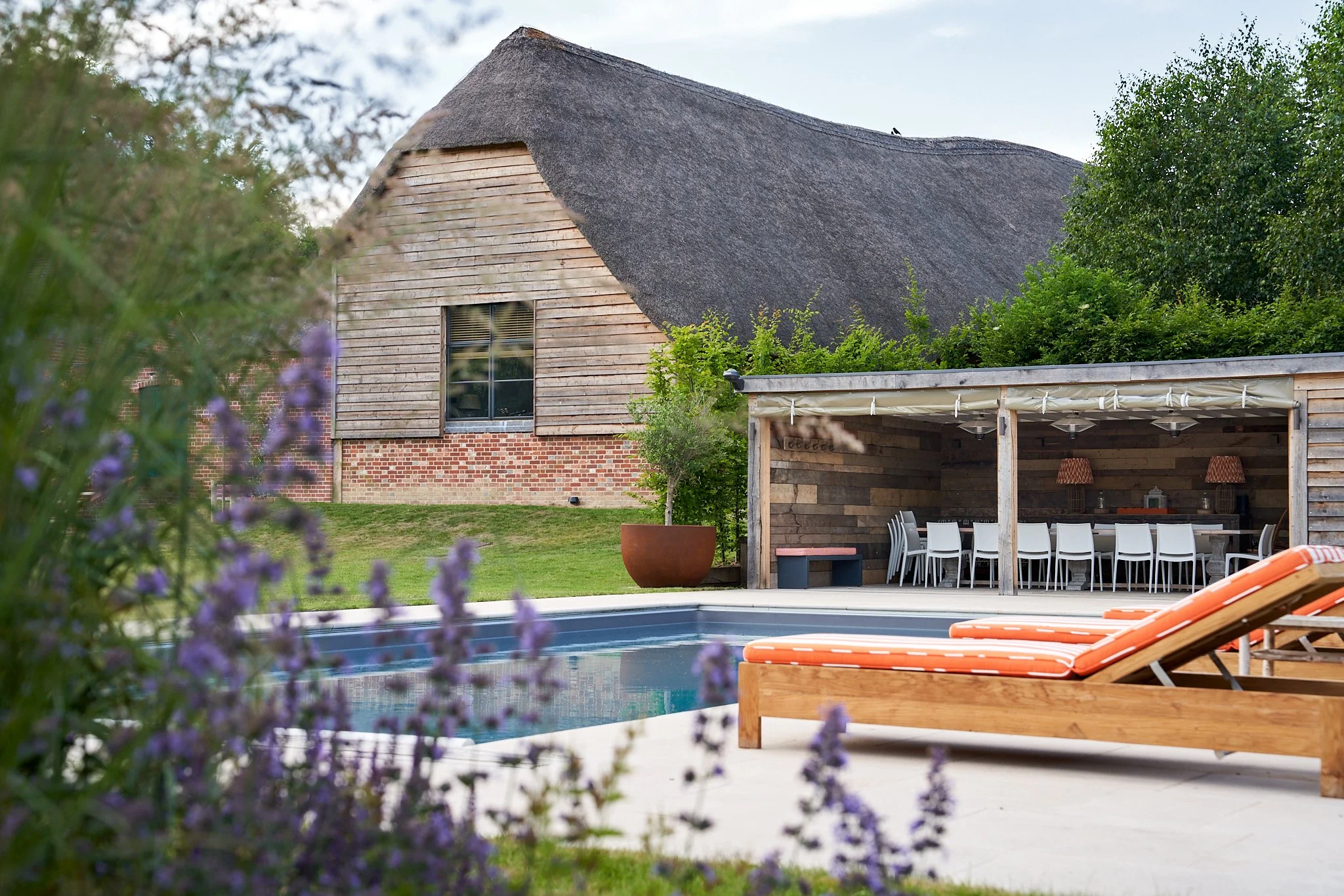Backyard with a swimming pool, orange and white lounge chairs, a covered outdoor dining area with white chairs, potted tree, and a rustic house with a thatched roof and wooden siding.