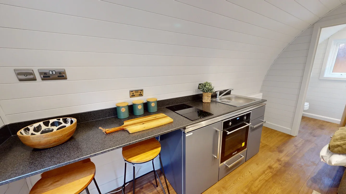 Small kitchen with white wooden paneled walls, black countertop, stainless steel stove and oven, small sink with a plant, and a window in the background. There are three teal canisters with yellow lids and a wooden bowl with black spots on the counter.