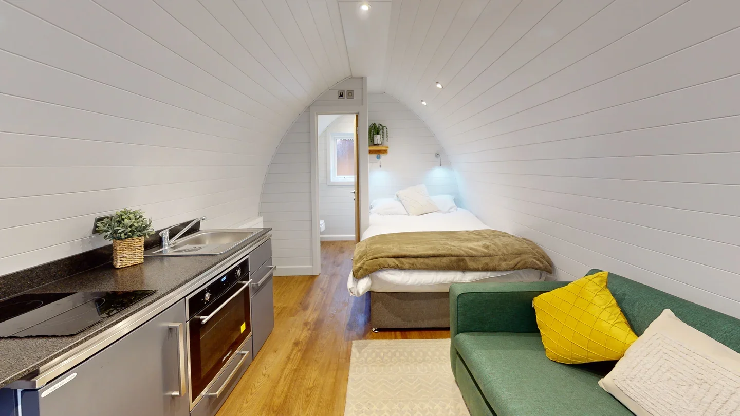 Interior of a small studio apartment with a kitchenette, a bed against the wall, and a green couch with pillows.